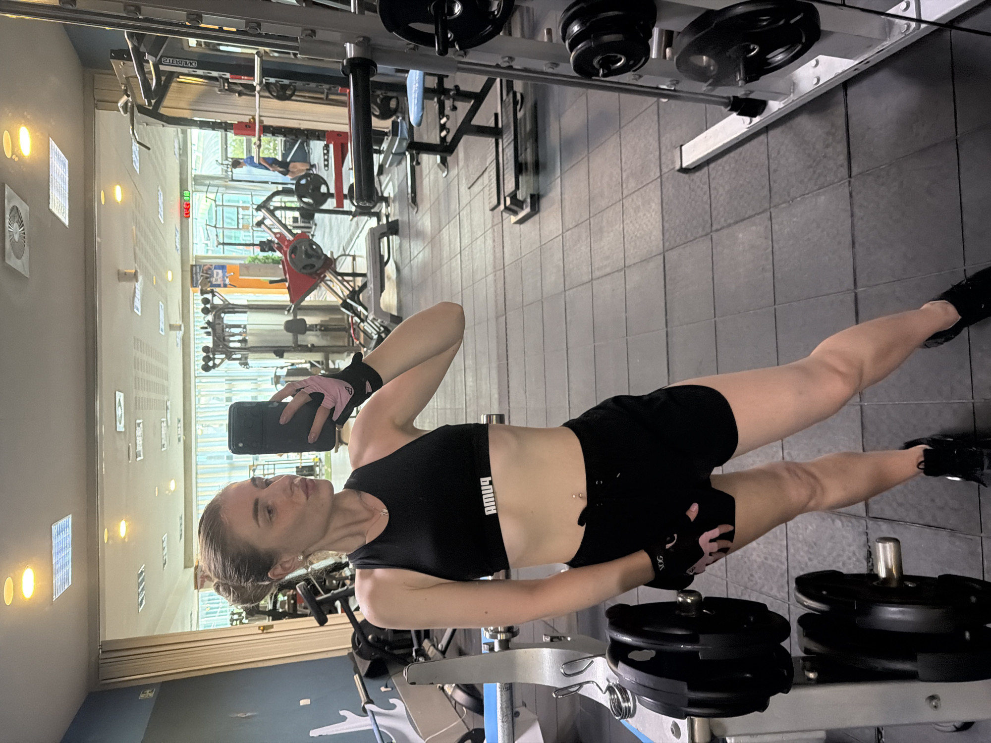 A woman in workout attire stands in a gym, taking a selfie. She is wearing a black sports bra, black shorts, and black sneakers. She has on pink and black weightlifting gloves. She has light brown hair pulled back in a bun. The gym has tiled floors and weightlifting equipment, including a smith machine, barbells, and weight plates. The lighting is artificial.