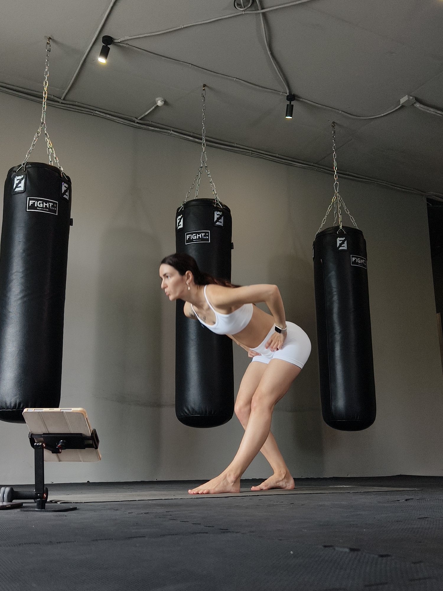 The image shows a woman in athletic wear performing a bent-over exercise in a gym with three punching bags hanging from the ceiling. The gym appears to be a boxing or martial arts studio. The woman is barefoot and focused on a tablet device on a stand in front of her. The punching bags are black with a white logo that says "FIGHT TECH". The walls are a neutral gray, and the floor is covered with black mats. The lighting is somewhat dim, with spotlights on the ceiling. The scene is devoid of any other people or activities. There is no information in the image to suggest the location is in Tyumen, Russia.