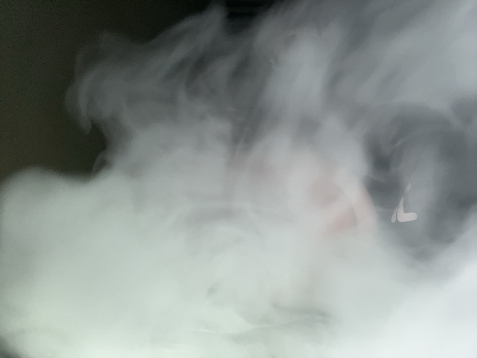 A forensic examination of the image reveals a partial human presence obscured by a dense, swirling cloud of white vapor or smoke. The scene is situated within an indoor or enclosed environment in the city of Chaikovsky, Russia. In the lower-center to right, a hand, displaying a reddish skin tone, is visible through the vapor. This hand is gripping a white, L-shaped object identified as the left side of a wireless earbud. The subject's face and upper body are largely indistinct due to the pervasive vapor. The background is uniformly dark, indicating a dimly lit setting. The substantial volume of vapor suggests an immediate prior action, such as exhalation from a vaping device.