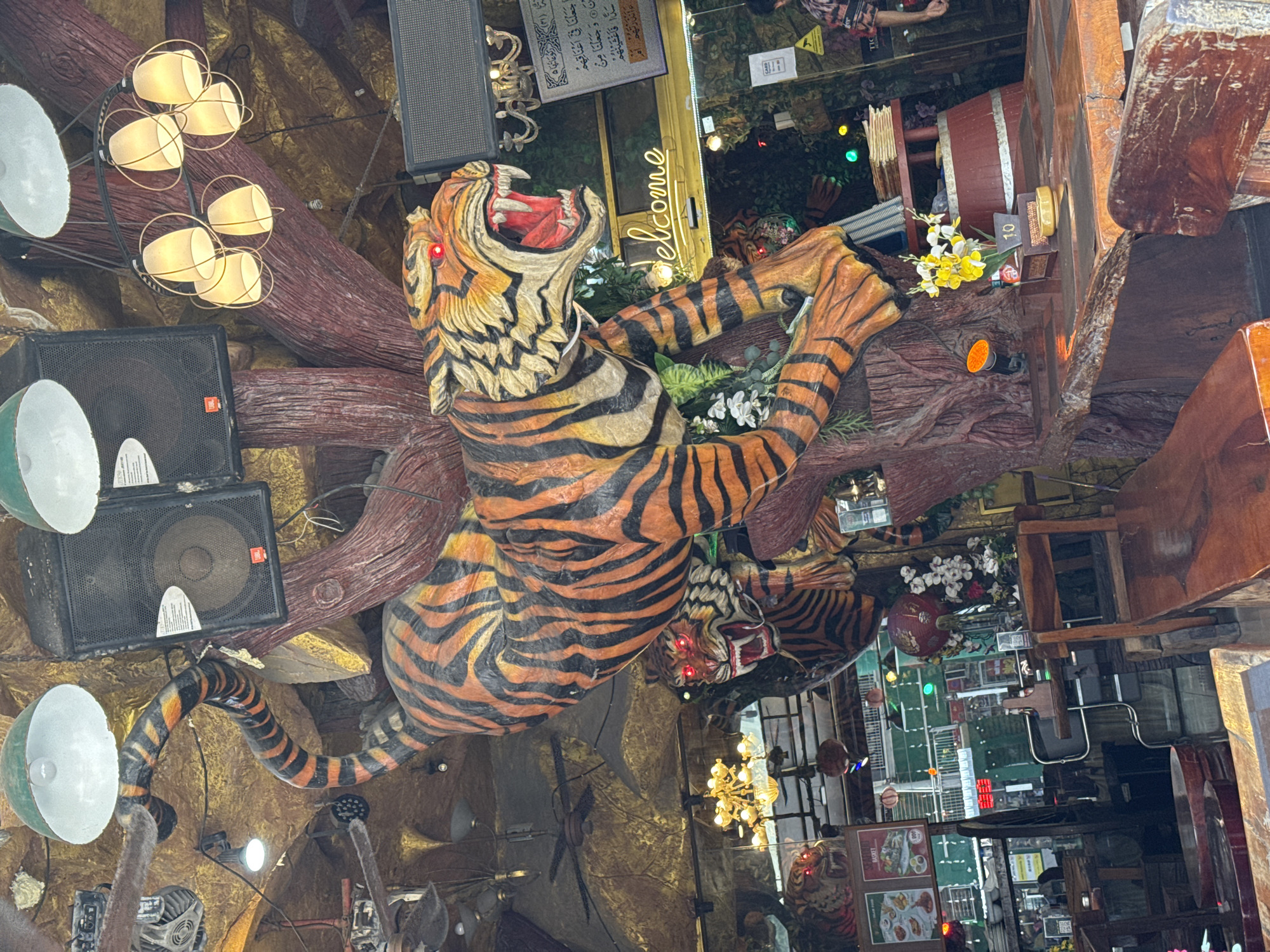 In Patong, Thailand, a restaurant interior features large, decorative tiger figures mounted on a faux tree structure. One tiger, predominantly orange with black stripes, is positioned leaping across the frame, its mouth open with visible fangs and red glowing eyes. A second, similar tiger head is visible below and to the left, also with glowing red eyes. The ceiling is adorned with multiple speakers, industrial-style fans, and a multi-bulb chandelier. Below the tigers, wooden tables and chairs are arranged. A neon sign reading "welcome" is mounted on a wall behind the main tiger figure. Further in the background, another sign with Arabic script is visible. The overall atmosphere appears to be themed, possibly jungle or animalistic, and is likely a lively establishment.