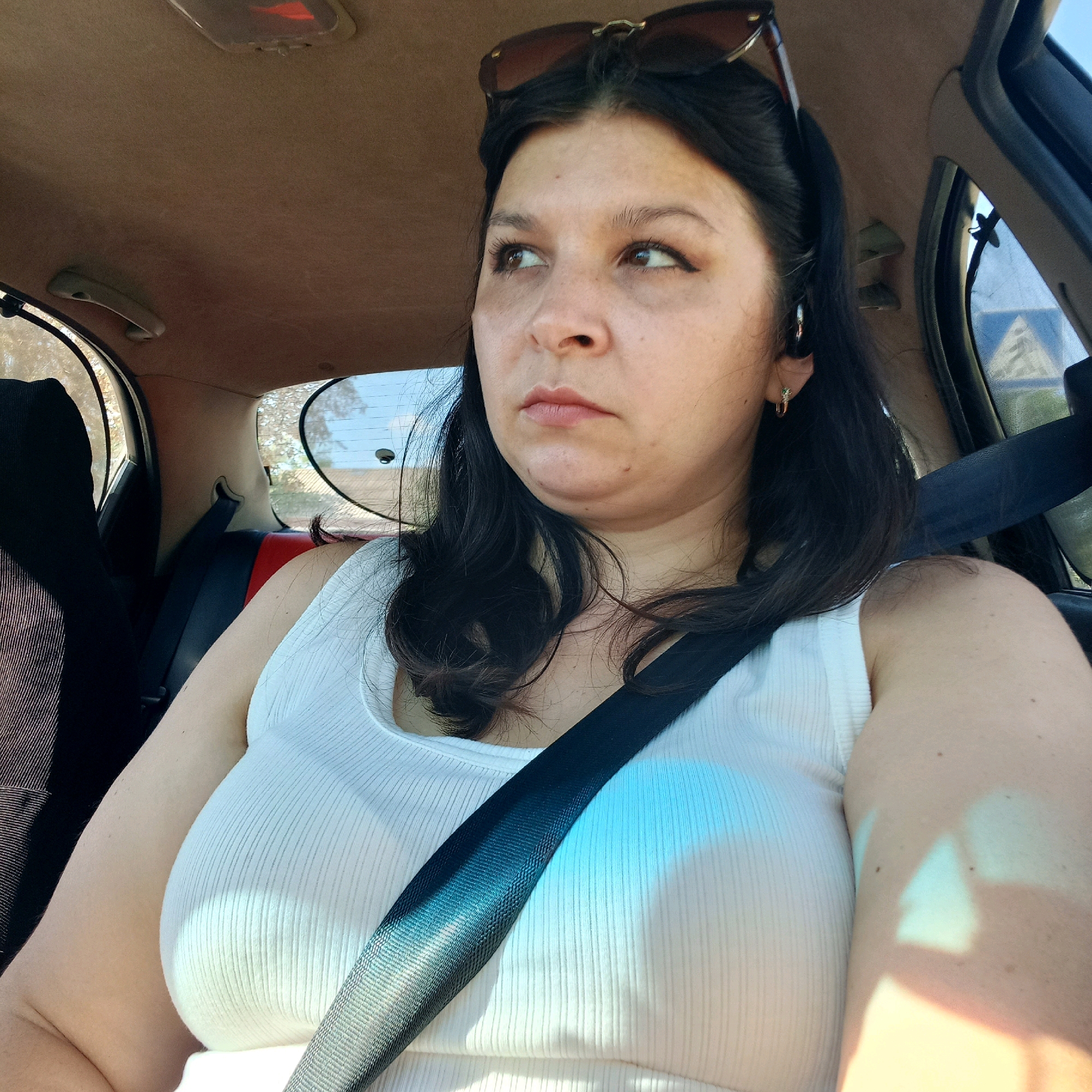 A medium shot shows a woman with dark hair and brown eyes sitting in the passenger seat of a car. She is wearing a white tank top and sunglasses are perched on her head. Her head is turned to the right, and she appears to be looking out the window. The interior of the car is visible, including the headliner, a seatbelt, and the passenger-side window. Outside the window, trees and a blue and white road sign can be seen. The lighting suggests it is daytime. The location specified, Klyntsi, Ukraine, is not evident from the visual information in the image.