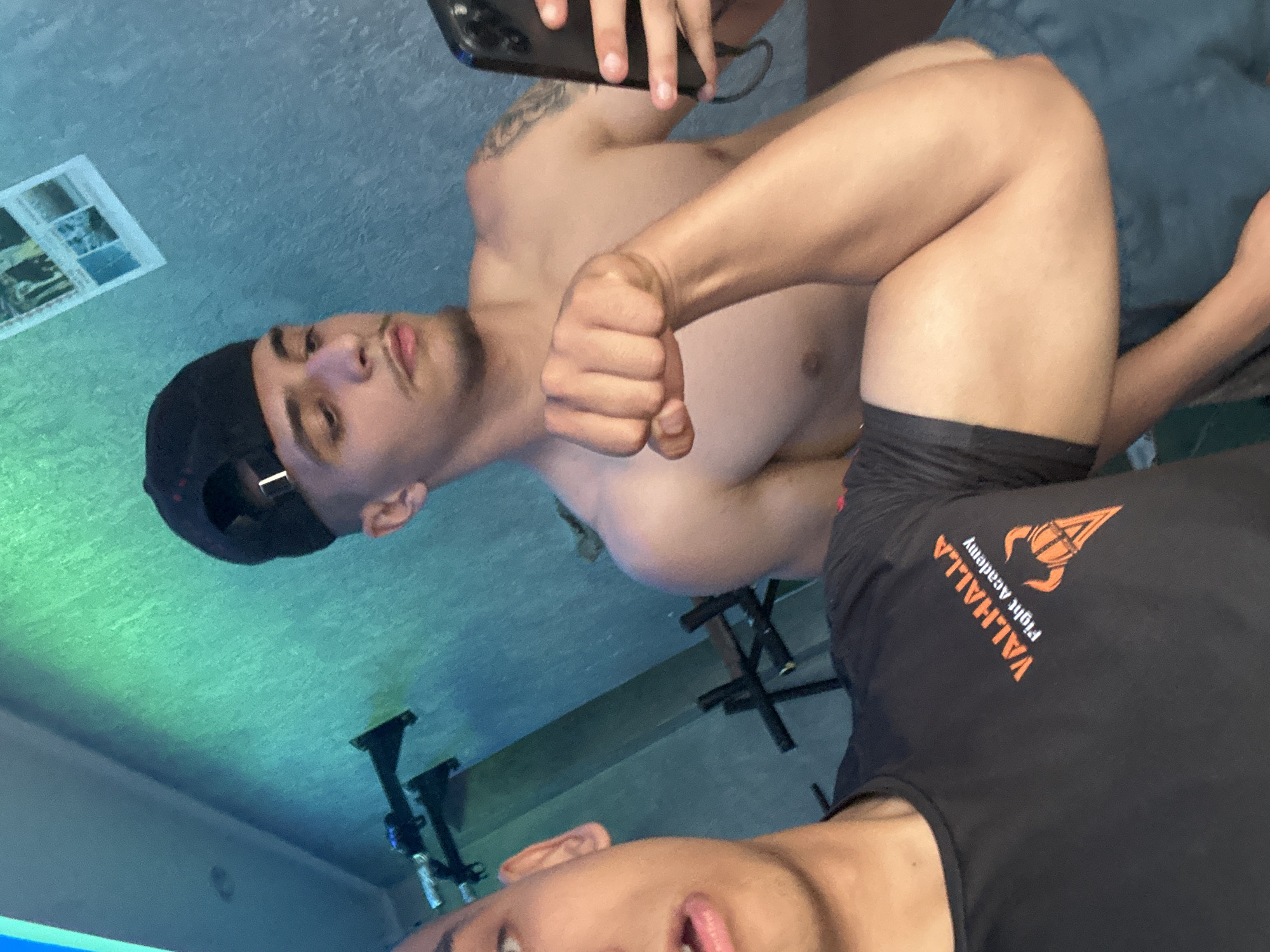 The image, rotated 90 degrees counter-clockwise, features two young males indoors, likely in a bedroom or personal space. The scene appears to be a casual selfie or photograph, possibly taken with a phone, which is partially visible in the upper right quadrant of the image.
In the background, a shirtless male with a lean build and a tattoo on his left shoulder/upper arm looks towards the camera. He wears a black baseball cap backwards. His right hand is clenched into a fist. He has short facial hair and his mouth is slightly open. Behind him, a dark blue bed or sofa is visible.
Partially visible in the foreground, another male looks towards the camera. He is wearing a black t-shirt with an orange logo and text on it. The text, which is upside down and rotated due to the image's orientation, reads "VALHALLA Flight Academy" with a graphic of a winged helmet or similar design.
The setting includes a light blue-green wall or ceiling, possibly indicating ambient lighting. A framed picture or collage hangs on the wall in the upper left. Dark, possibly metallic, furniture or equipment can be seen in the lower left and center background. The overall mood appears neutral. The time of day is not discernible due to indoor lighting.
