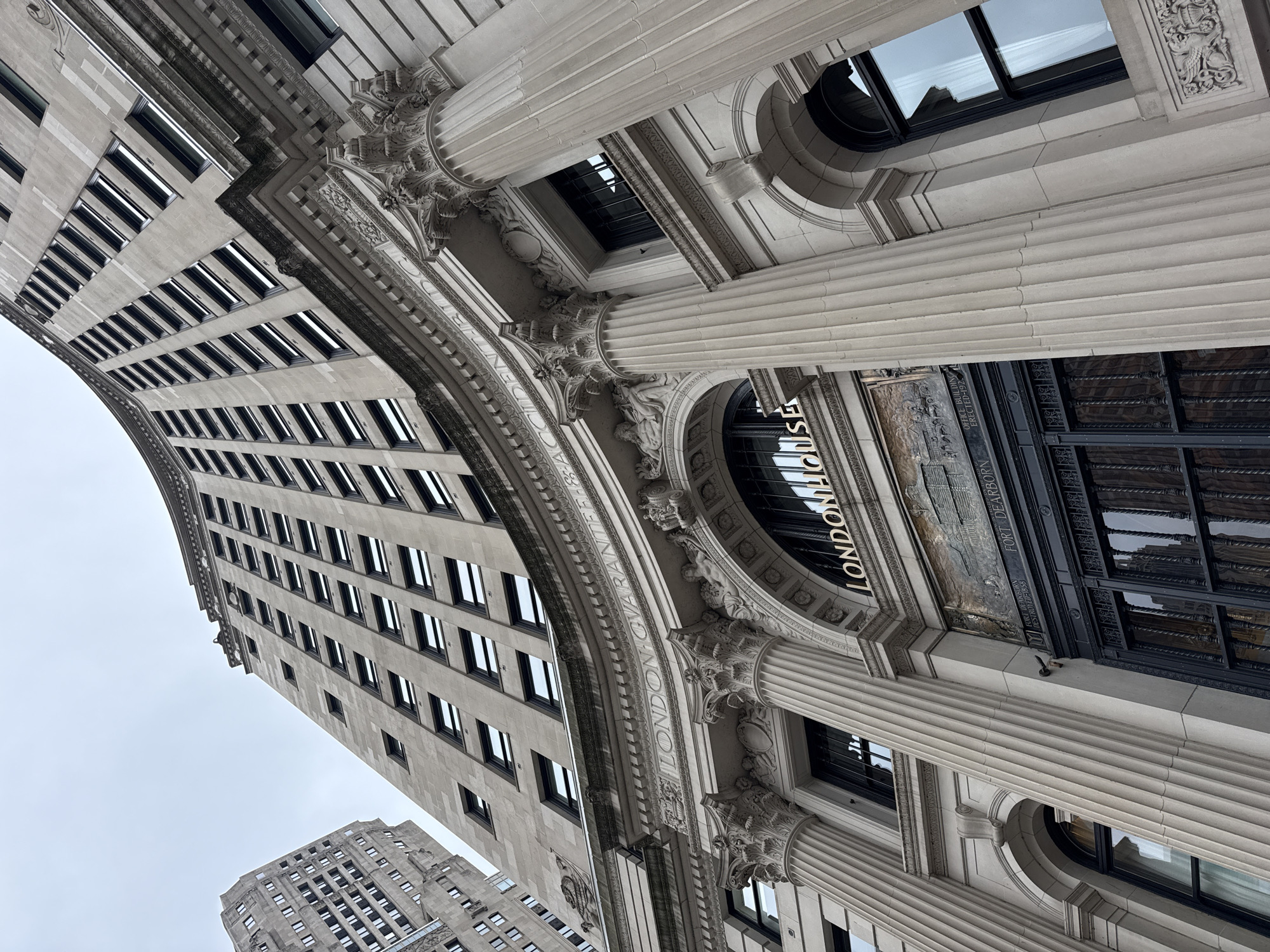 This media file captures an upward-angle view of a grand, classical-style building in Chicago, USA, likely on an overcast day. The architecture is characterized by ornate stone carvings, fluted columns, and a curved facade lined with numerous windows. The building's facade displays the engraved text "LONDON GUARANTEE" and "FORT DEARBORN" as well as "OFFICE BUILDING ERECTED 1921". The image focuses on the intricate details of the building's upper levels, including decorative cornices and archways. In the background, another, taller building with a similar architectural style is visible, suggesting an urban setting with a concentration of historic architecture. The overall impression is one of architectural grandeur and historical significance.