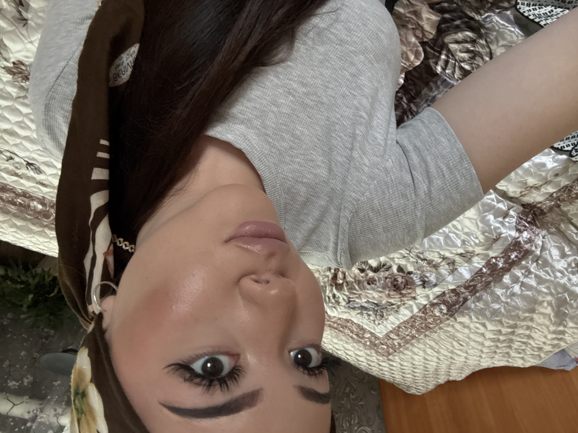 A selfie photograph shows a woman's face from a low angle. The woman has dark brown hair and is wearing a gray, short-sleeved shirt. A brown headscarf with white patterns is partially covering her hair. She has fair skin, dark eyebrows, and eyelashes, and her lips appear to have a light gloss. She is wearing a silver hoop earring on her left ear.
The background consists of a patterned, quilted fabric with beige, white, and brown tones. Part of a wooden surface is visible in the lower-right corner of the image.
There is no information available to determine the location of the image within the city of Konovalovka, Russia.