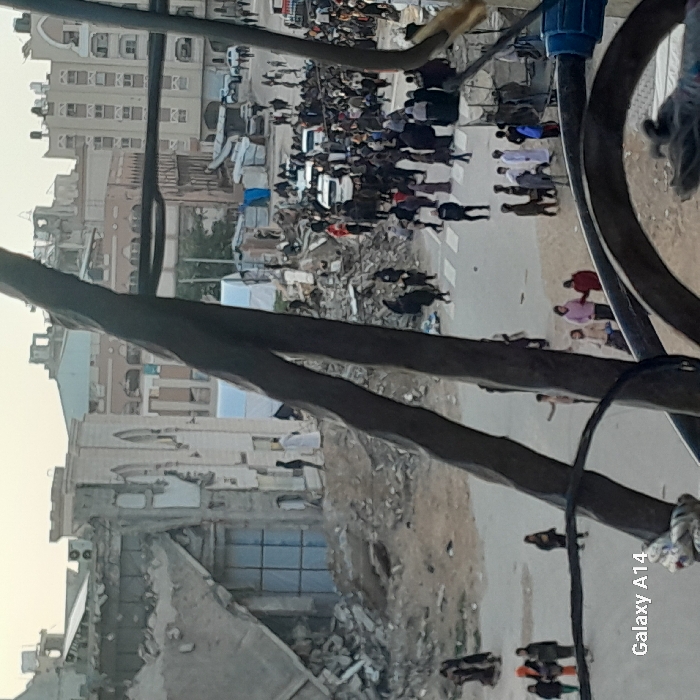 The image depicts a crowded, damaged urban street scene in Khan Yunis, Palestinian Territory, viewed through dark, out-of-focus metallic structures in the immediate foreground.
In the mid-ground left, a large, partially collapsed building with distinctive arched window frames stands amidst significant piles of concrete rubble and debris. A prominent section of a large doorway or window on this building's ground level is covered with a light blue tarp or material. Two individuals in white thobes are visible near the base of this damaged structure, standing on disturbed earth.
The central and right mid-ground is dominated by a wide, dusty street or open area filled with numerous individuals, primarily adults and some children, walking and congregating. Many women are wearing hijabs or niqabs, and men are dressed in a mix of traditional and casual attire. Two white civilian vehicles are visible within the crowd, appearing to navigate or be stationary amongst the people. The immediate edges of the street show further debris and disturbed ground.
In the background, beyond the main crowd, additional makeshift shelters or tents, some white, some blue, are erected amidst more rubble. Farther back, a dense urban landscape of multi-story buildings, some appearing damaged or partially destroyed, extends to the horizon. Rooftop water tanks are visible on several buildings. The sky is uniformly pale and overcast. The bottom left corner of the image contains a "Galaxy A14" watermark.