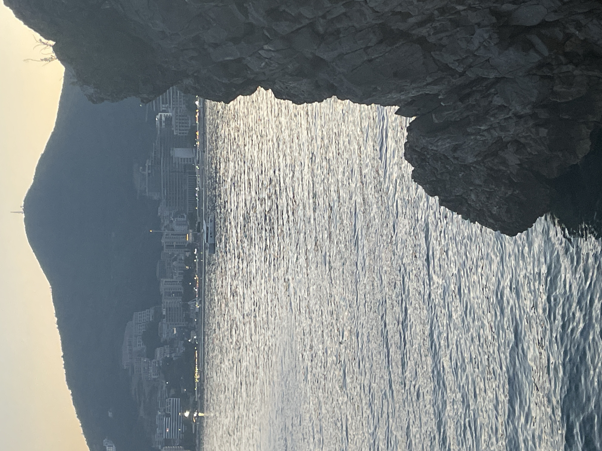 A low-angle shot shows a tranquil bay and cityscape as seen from a cave or rocky overhang on a sunny day. The glistening water takes up most of the scene, with the city skyline just visible along the horizon. The cityscape comprises high-rise apartments and hotels, some with lights on, indicating that it might be early morning or late evening. A dark mountain stretches along the left, silhouetted against the light sky. A rocky overhang frames the view at the top of the image, with a section of a cave ceiling and a rugged edge that leads into the sea. It is likely the Mogren beach area in Budva, Montenegro.