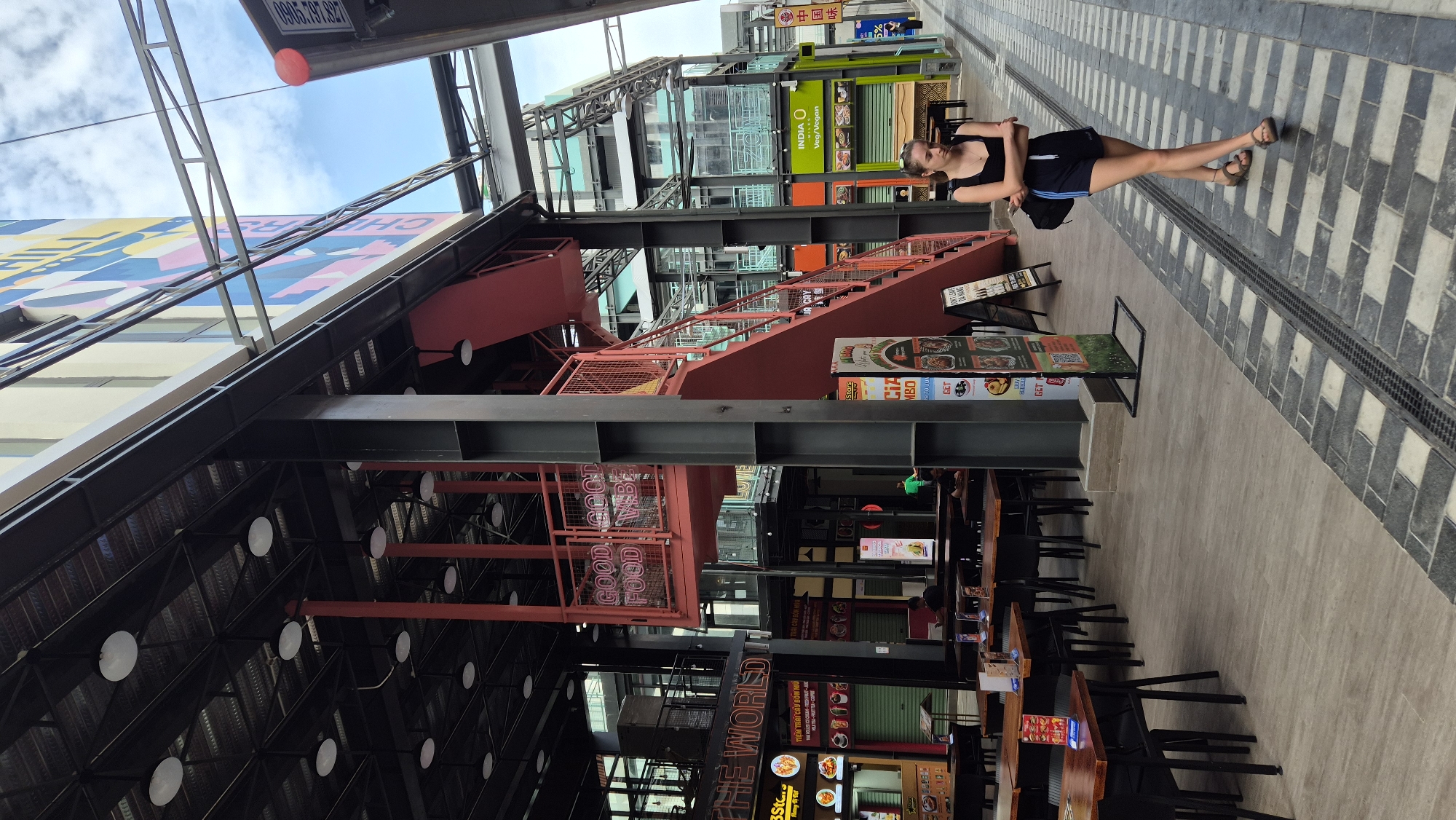The image depicts an outdoor dining area and walkway in Đà Nẵng, Vietnam. A young woman wearing a black tank top and dark shorts with blue stripes is walking away from the camera on a patterned tiled walkway. She has her arms crossed and is wearing sandals. To her left, there is an open-air structure with exposed metal beams and a red staircase leading up to a mezzanine level. The mezzanine has a red railing and displays neon signs that read "GOOD FOOD GOOD VIBES". Below the mezzanine, there are several tables and chairs set up for dining, some with umbrellas. Various food stalls and shops are visible in the background, some with closed green metal shutters. One stall has a sign with Vietnamese text and images of food. The overall scene suggests a modern urban environment with a focus on food and leisure.