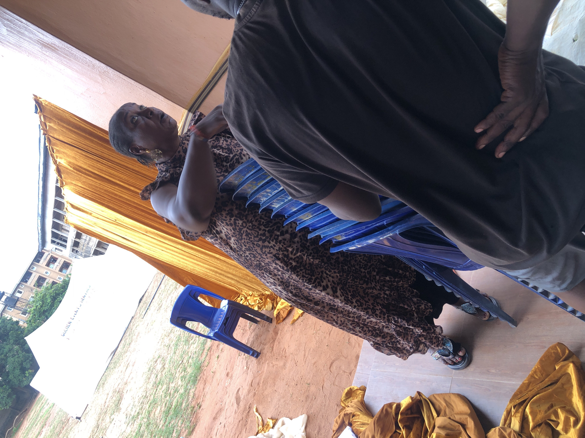 The image, likely taken in Onitsha, Nigeria, shows two individuals. A woman in a leopard-print dress is standing with her arms crossed, looking upwards. She is wearing gold earrings and sandals with bows. She is positioned next to a stack of blue plastic chairs. A man, partially visible, is standing beside her, wearing a brown t-shirt and light gray shorts. His hand is on his hip. A single blue plastic chair is situated on a dirt patch near a white tent with "GOLDEN EVENTS RENTALS" printed on it. Golden fabric is also draped on the ground. In the background, there is a building with multiple apartments. The ground is a mix of dirt and grass.