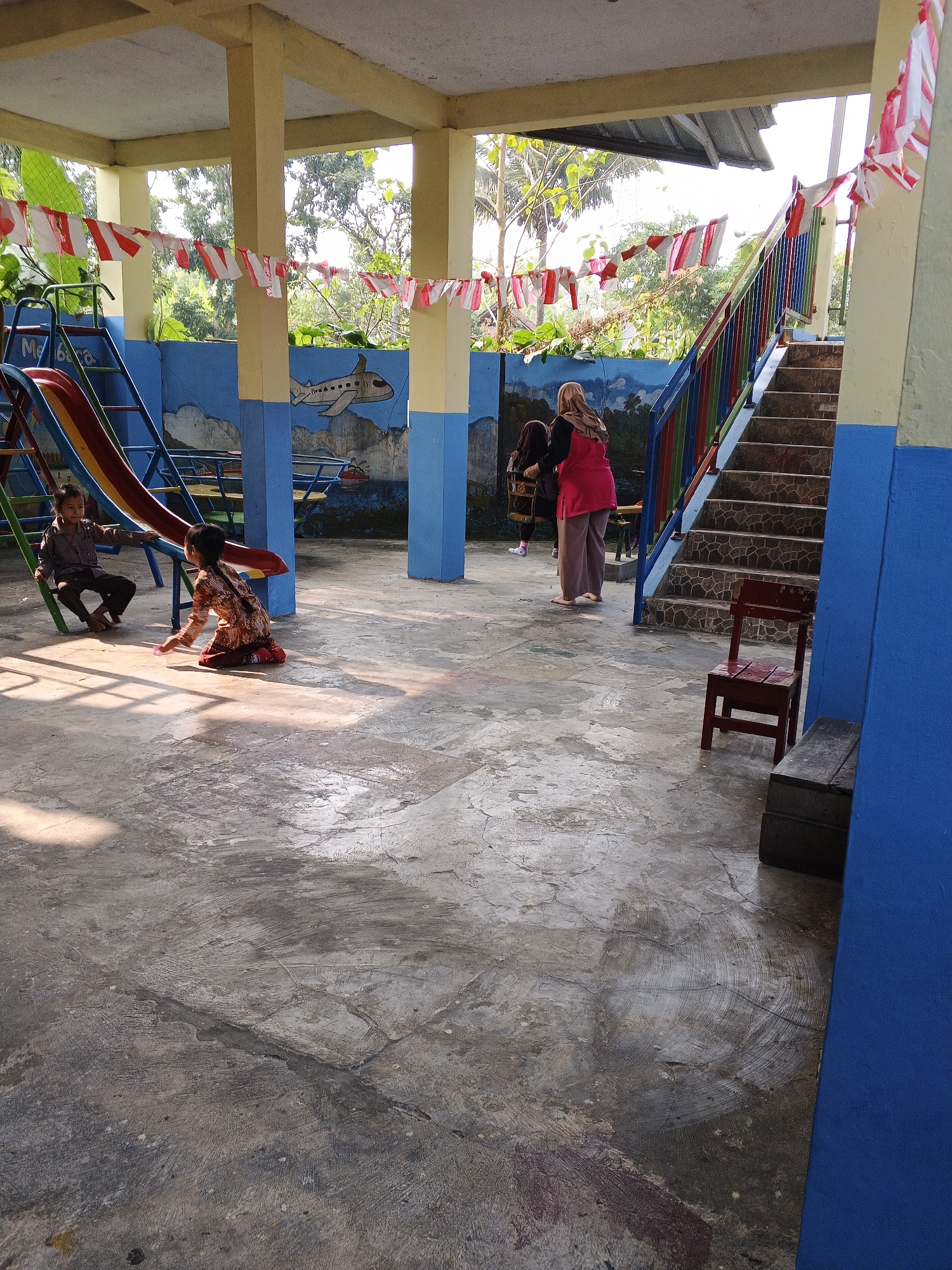 In Warungkiara, Indonesia, an outdoor covered area with a concrete floor is depicted. Red, white, and blue flags are strung across the structure. A blue wall features a mural of an airplane and clouds. In the foreground, two children are playing. One child, wearing a brown uniform, sits on a playground slide. Another child, wearing a patterned shirt and pants, kneels on the ground. To the right, a staircase with a colorful railing leads up to an unknown area. A red wooden chair and a dark wooden box are placed near the stairs. Two women, one wearing a pink headscarf and vest over a black shirt and pink pants, and another in a dark headscarf and brown clothing, are standing near the stairs.
