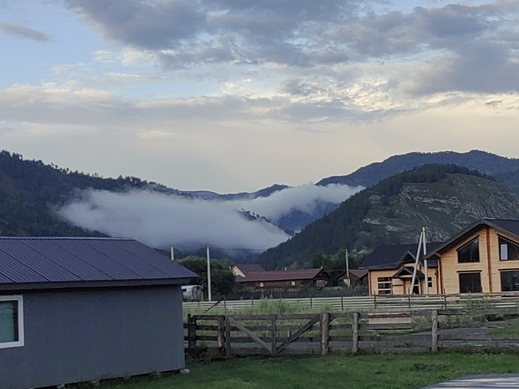 The image depicts a rural landscape with mountains in the background. A thick band of fog or low clouds obscures the mid-section of the mountains, creating a distinct visual separation between the lower, forested slopes and the higher peaks. The sky above is partly cloudy, with a mix of light and dark gray clouds against a pale blue sky.
In the foreground, there are several buildings and elements of a rural settlement. To the left, a dark gray building with a dark, corrugated metal roof is visible, featuring a single white-framed window. In the center and to the right, there are wooden structures, including a fence with a gate made of rustic timber. Behind the fence, there are houses with wooden exteriors and dark roofs. One prominent house on the right is a large, modern wooden structure with a distinctive A-frame design and large windows. Another longer, traditional wooden building with a red roof is visible further back. The ground in the foreground is covered in green grass. The scene appears to be set in Anos, Russia, a region known for its mountainous terrain and natural beauty, often associated with dacha (summer house) settlements.