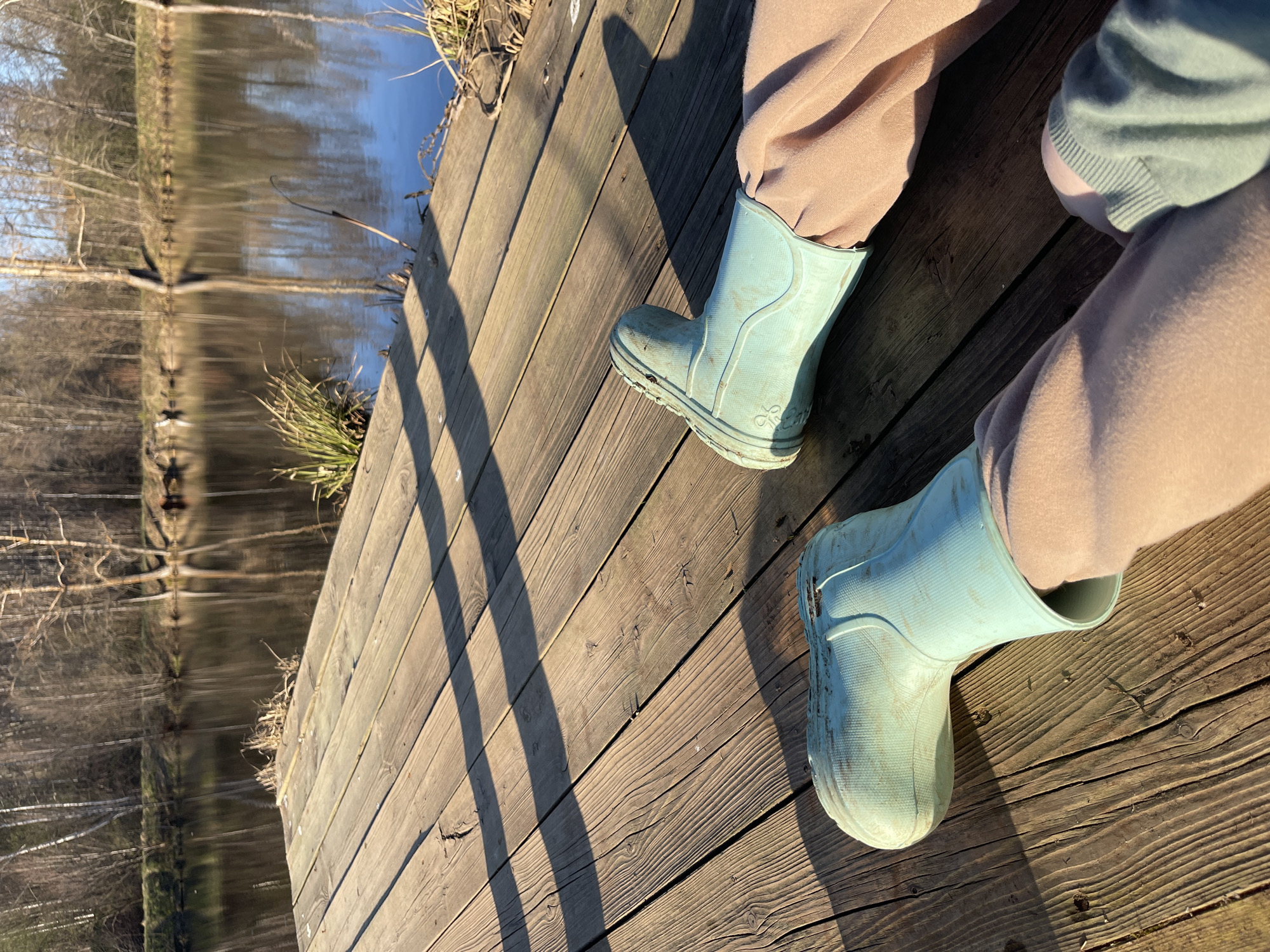The image shows a person sitting on a wooden deck next to a body of water, possibly a lake or pond. The person is wearing tan-colored sweatpants and light blue rain boots. The wooden deck is made of planks and there are shadows cast across it. The water reflects the trees on the opposite bank. The environment seems natural, suggesting a park or a rural area. The city of Zhigalki, Russia is not directly discernible within the frame, so further information would be needed to definitively say this photo was taken there.