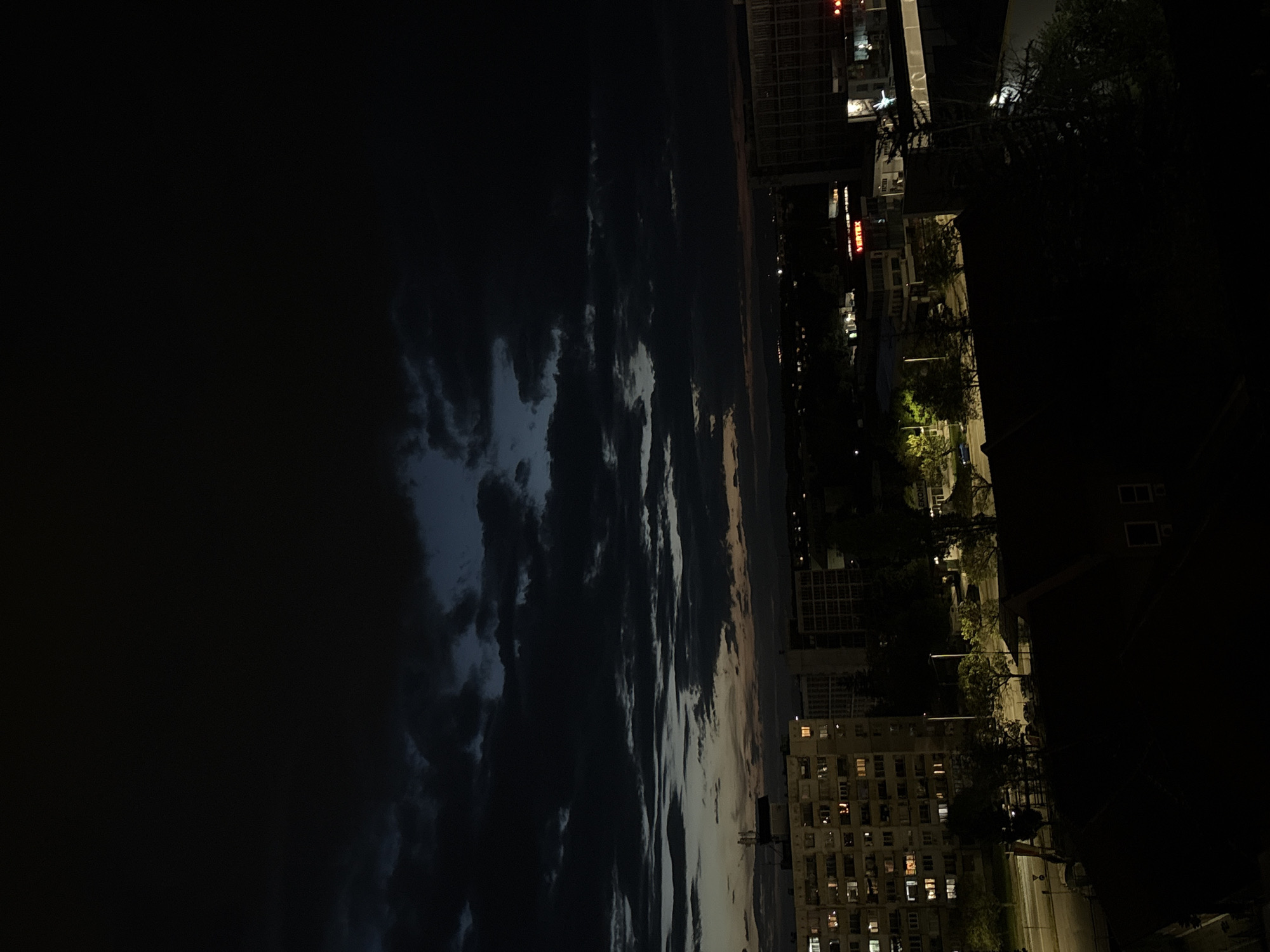 The image depicts a nighttime cityscape in Sofia, Bulgaria, with the sky dominating the left side of the frame. The sky shows a mix of dark clouds and patches of light, likely illuminated by the setting sun or city lights. On the right, buildings are visible, some with illuminated windows. Streetlights are also present, casting light on the streets and surrounding trees. The scene suggests a residential area near a park or open space, with the city's illuminated structures contrasting against the darkening sky.