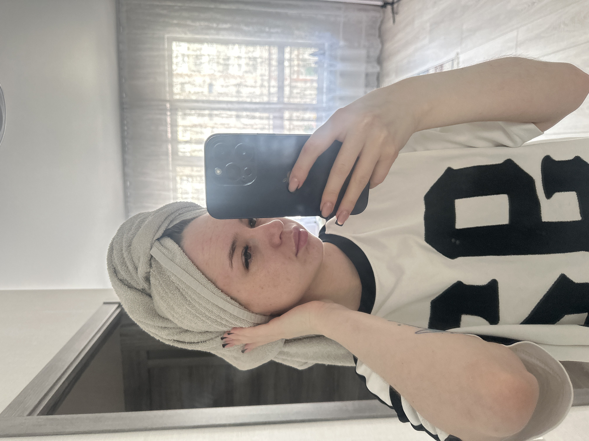 A young woman is depicted taking a mirror selfie, holding a black smartphone with her right hand. Her head is tilted to the left, resting gently on her left hand, which is wrapped in a light grey towel. She has fair skin with visible freckles and a soft, slightly pensive expression with her lips subtly parted. Her right hand displays light-colored nails with black tips, while her left hand has darker polish. She is wearing a white short-sleeved t-shirt with a black trim around the neckline and sleeves, and a prominent black number "89" is printed on the front. The background reflects a window with light-colored blinds or curtains, suggesting an indoor setting during daylight hours. A light-colored wall and what appears to be a mirror frame or counter are also visible in the reflection. A faint tattoo can be seen on her left arm.
