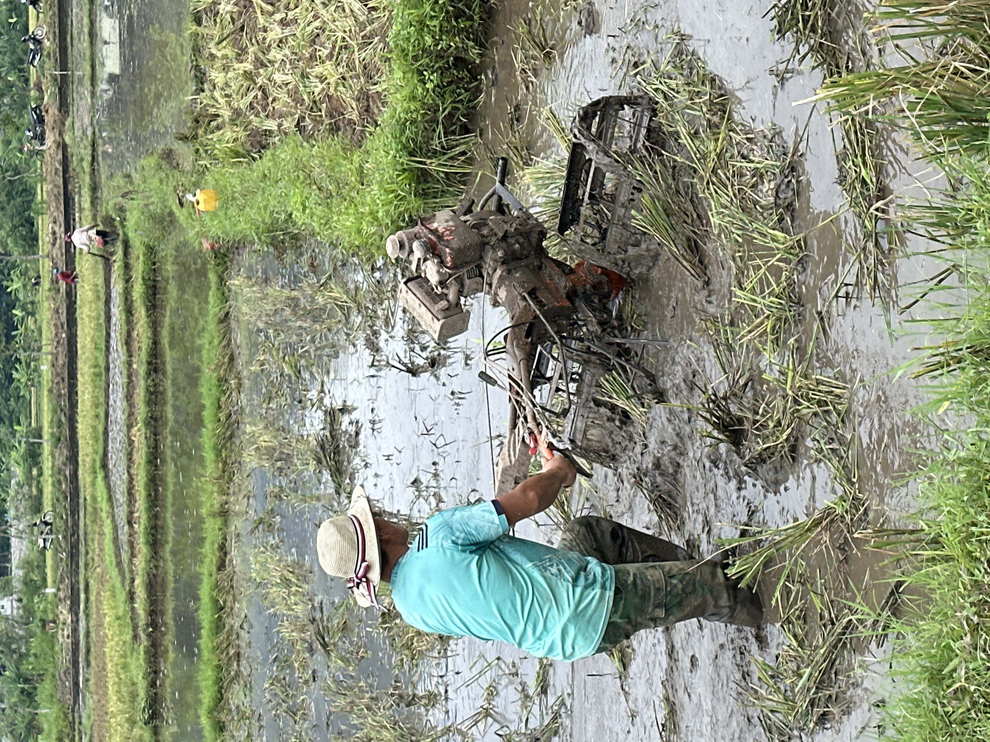 A person wearing a straw hat and a teal shirt is operating a tiller in a muddy rice paddy. The person is facing away from the camera, pushing the tiller. The tiller is red and orange and covered in mud. The rice paddy is flooded and filled with cut rice stalks. In the background, there are more rice paddies, some with water and some dry. There are also several motorcycles parked on a path in the distance, and a person in a red shirt is working in another rice paddy. The scene appears to be in Mai Chau, Vietnam, given the typical agricultural landscape of the region.