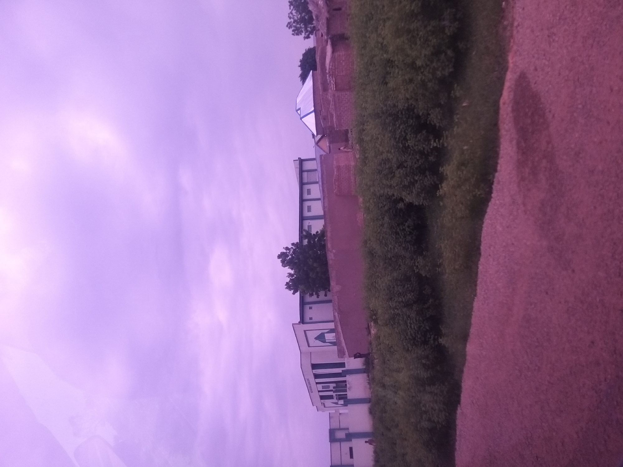 The image depicts a street view in Dukku, Nigeria, under a purple-tinted sky. In the background, a modern building with white walls and dark windows stands prominently, featuring a green geometric design on its facade. To the right of this structure, there are buildings constructed with reddish bricks, some with thatched roofs and one with a silver, pitched roof. A dense row of green bushes and trees separates the foreground from the buildings. The foreground consists of a gravel road with a reddish hue. A single person, appearing as a dark silhouette, is visible near the entrance of the brick structures, and another figure is seen walking near the white building. The overall scene suggests a developing area with a mix of modern and traditional architecture.