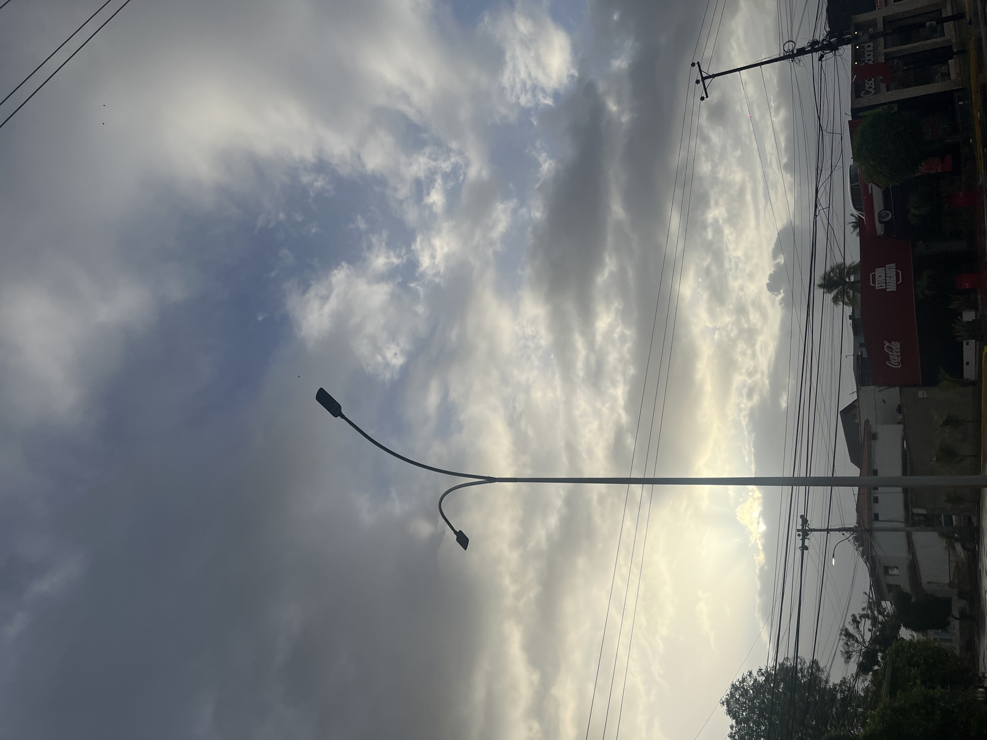 The image shows a sky with many clouds. A streetlight is positioned in the foreground, and multiple utility poles with overhead wires are present along the right side of the frame. Buildings are visible on the right, with a Coca-Cola sign prominently displayed on one of them. The scene appears to be in an urban area, potentially Pampatar, Venezuela. Vegetation is visible at the bottom right.