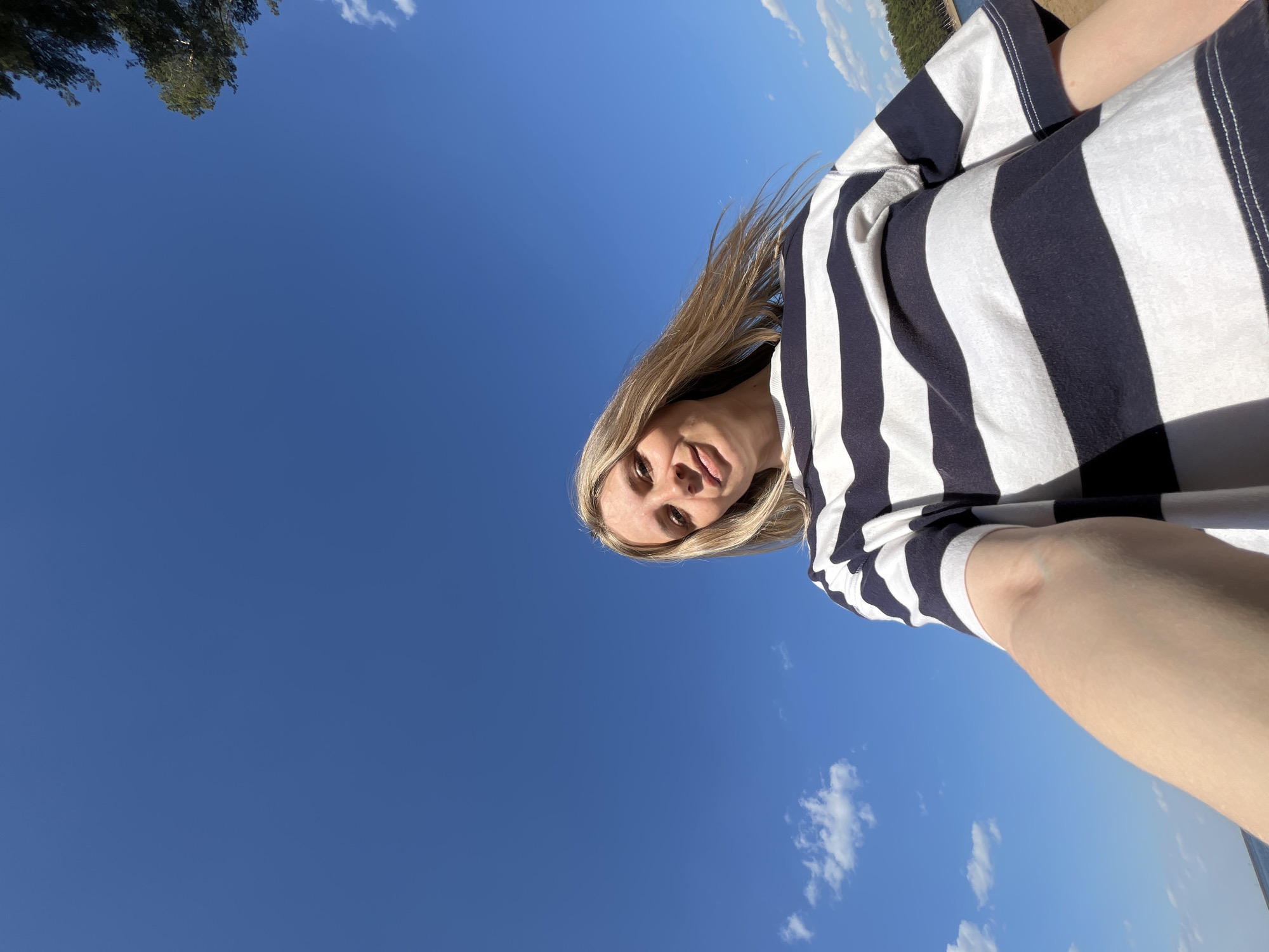 A low-angle selfie taken outdoors under a clear blue sky, likely in the city of Chaikovsky, Russia, features a young woman with blonde hair wearing a striped black and white shirt. The sky is partially visible, with a few scattered white clouds. A glimpse of a tree can be seen in the upper left corner of the frame, while a body of water or a landscape is faintly visible along the top edge towards the right. The woman's face is angled towards the camera, with visible light skin.