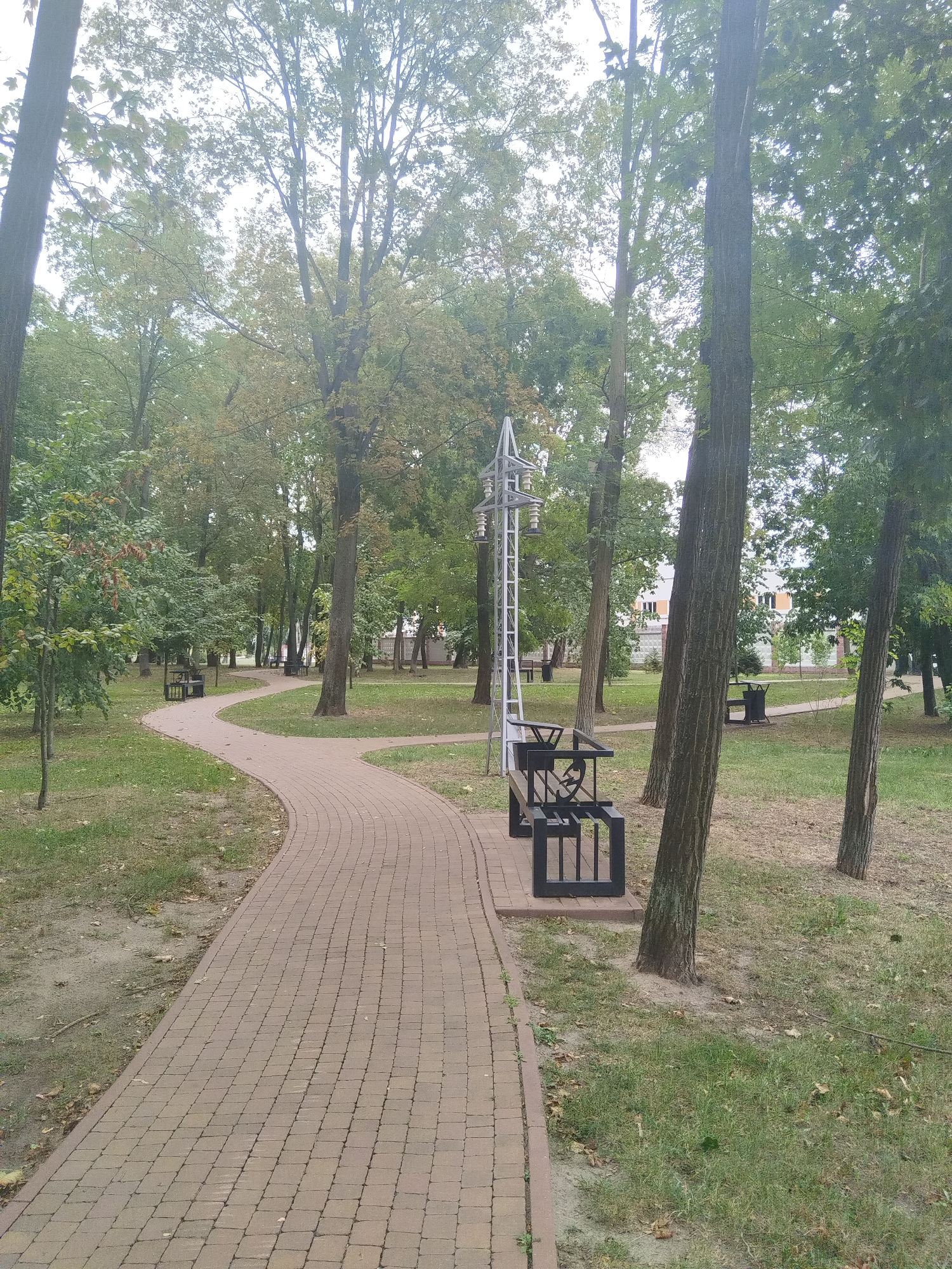 The image depicts a park path in Brest, Belarus. A curved, brick-paved walkway winds through a grassy area dotted with trees. A black metal bench with a decorative backrest sits alongside the path. In the background, more trees are visible, as well as a building with white walls and windows. The sky is overcast.