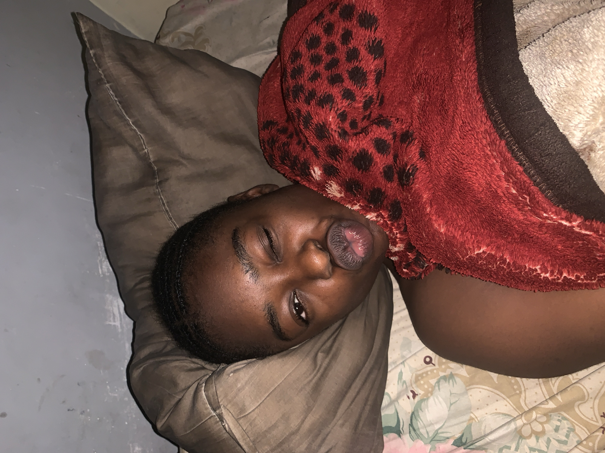A Black woman lies on a bed. Her head rests on a taupe pillow with a visible seam. A red, fuzzy blanket with black leopard spots covers part of her head and body. The bed has a floral-patterned sheet. The woman's eyes are partially closed, one appears to be winking, and her mouth is slightly open. The setting appears to be indoors, potentially a bedroom. There is no indication of the location being Rafin Par, Nigeria.