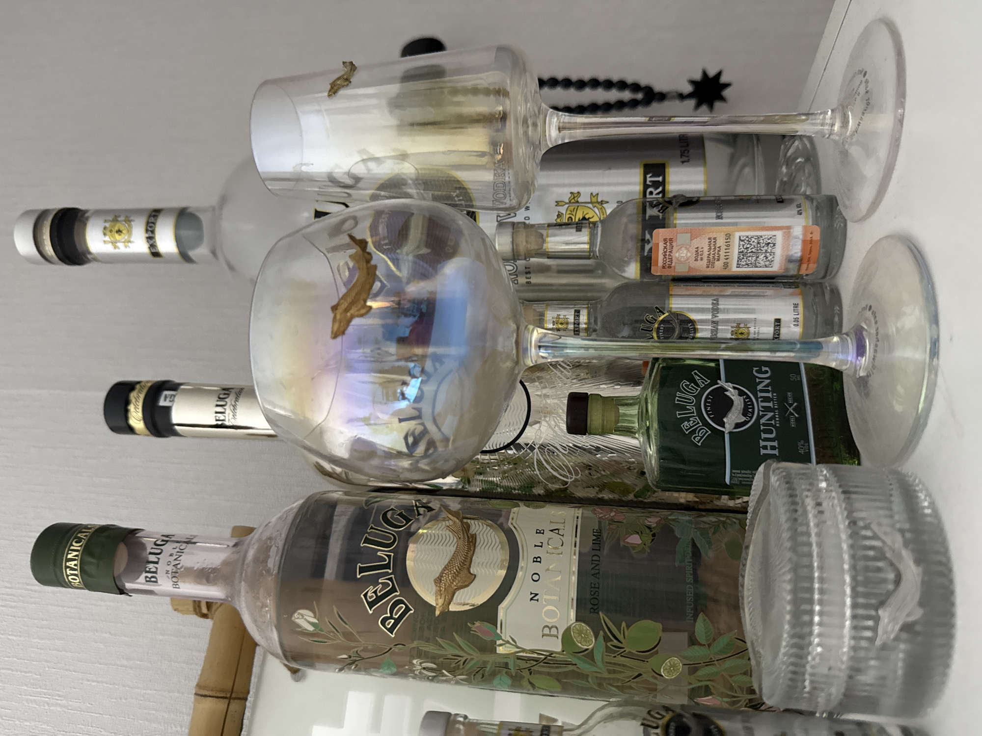 A collection of Beluga vodka and spirits bottles, along with associated glassware and decorative items, is meticulously arranged on a flat white surface against a light-colored, subtly textured wall in Vniissok, Russia.
Dominating the left-center is a large bottle of Beluga Noble Botanicals Rose and Lime infused spirit drink, identifiable by its dark label with gold text, a prominent gold sturgeon emblem, and a green cap inscribed "BOTANICALS." Partially obscured behind it, to the right, is another Beluga bottle with a black cap and gold "Celebration" inscription. In the upper right background, a large 1.75-liter bottle of Beluga Export Vodka is visible, featuring a white cap, silver band, and gold crest.
In the foreground, two stemmed glasses are positioned. The left glass displays an iridescent finish and a gold sturgeon emblem on its bowl, while the right glass is clear with a subtle golden tint and a smaller gold sturgeon emblem. Both glasses have clear stems and bases, with "BELUGA" etched into their undersides.
Further right, a small green 50 ml bottle of Beluga Hunting Herbal Bitter stands beside a 0.2-liter bottle of Beluga Vodka. This smaller vodka bottle features an orange excise stamp marked "РОССИЙСКАЯ ФЕДЕРАЦИЯ" (Russian Federation) and a QR code, indicating a 40% alcohol volume. On the far left, a 0.05-liter bottle of Beluga Export Vodka is visible next to a clear, ribbed cylindrical container with a sturgeon emblem embedded in its side.
Decorative elements include a horizontal bamboo pole in the upper-left background and a black beaded necklace with a black star charm hanging on the far right. The scene is brightly and evenly lit, presenting a static display of these items.