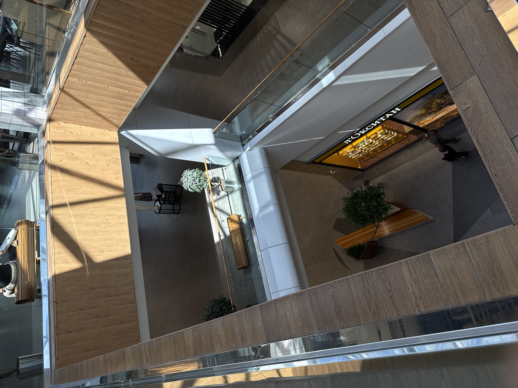 The image shows an interior view of a modern multi-level shopping mall, likely in Izhevsk, Russia, given the Cyrillic signage. The perspective is from an upper level looking down into the atrium and on the floor below. The architecture features clean lines, natural wood paneling on some walls and railings, and glass barriers along the walkways and staircases. Sunlight streams in from above, casting distinct shadows across the wooden surfaces and floors, suggesting it is daytime.
On the upper level visible, a wooden bench with a curved design is present, with a person seated on it. In the mid-level atrium, more wooden benches and potted plants are strategically placed, creating a relaxed atmosphere. A shop with bright yellow signage displaying Cyrillic text, which appears to read "ЛОКСИТАН" (likely a store name, possibly "L'OCCITANE"), is prominently featured on the lower floor. Two individuals are visible near the entrance of this shop, one of whom appears to be reaching out towards the other. The flooring in the mall is a neutral tone, likely tile or polished concrete, contributing to the overall modern aesthetic. The scene depicts a typical commercial environment, with elements of design and natural light contributing to its ambiance.