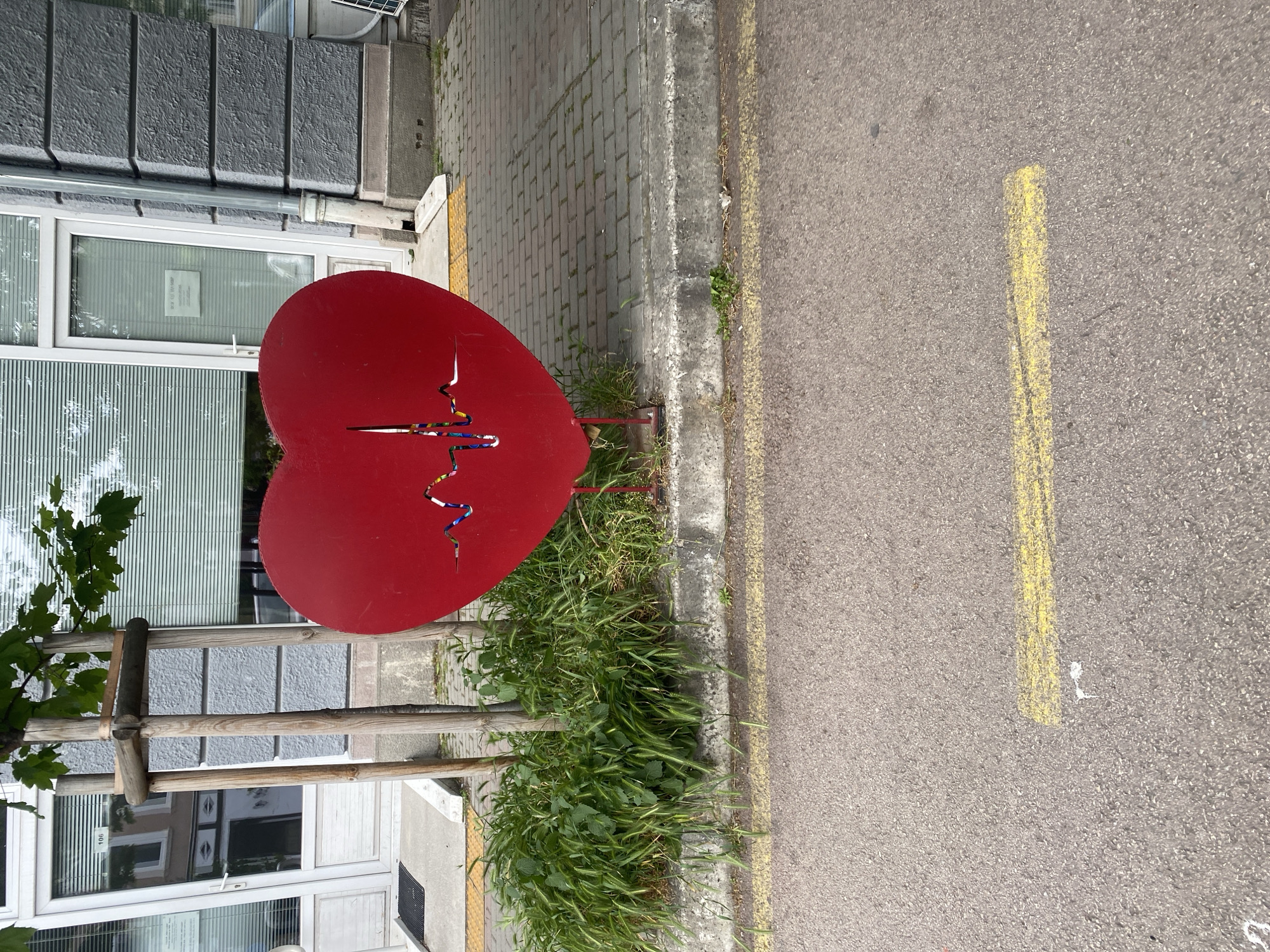 A red, heart-shaped metal sculpture stands on the sidewalk next to a street in Varna, Bulgaria. The heart is decorated with a colorful, stylized line. A single, vertical cut is visible on the heart. The sculpture is positioned next to a building with a gray stone exterior, windows with white frames, and horizontal blinds. Grass and weeds grow around the base of the sculpture and along the curb of the sidewalk. A yellow line is painted on the asphalt street next to the sidewalk. A small tree is visible to the left of the heart. The image is taken during daylight hours with natural lighting.