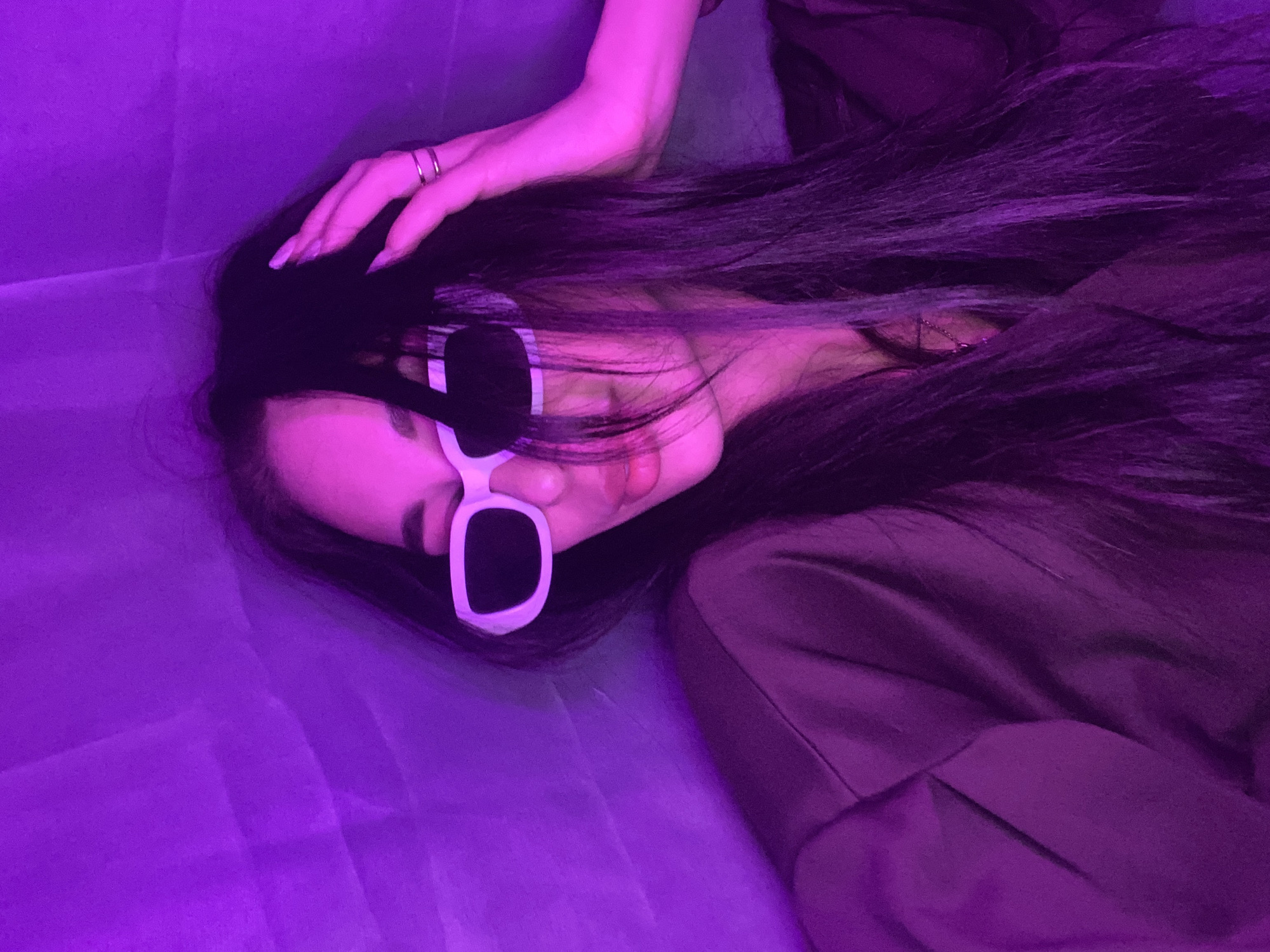 A woman with long, dark hair and white-rimmed sunglasses lies on a purple-toned surface. She has a ring on her finger. The scene is illuminated by a strong purple light, making it difficult to discern specific details of the environment. There is a piece of brown clothing around her shoulder. The location is unknown.
