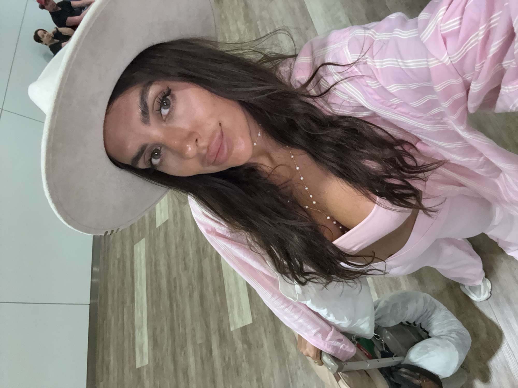 An eye-level, close-up, selfie-style shot captures a woman wearing a wide-brimmed hat and a pink and white striped shirt. She is holding onto the handle of a suitcase. The background is blurred, showing a light-colored wall and a wooden floor. Two people are visible in the background, sitting and facing away from the camera. The woman is the primary subject, with her face and upper body dominating the frame. She has dark hair and is wearing a necklace. The lighting is even, with no harsh shadows. The image appears to be taken indoors, possibly in an airport or a public waiting area.