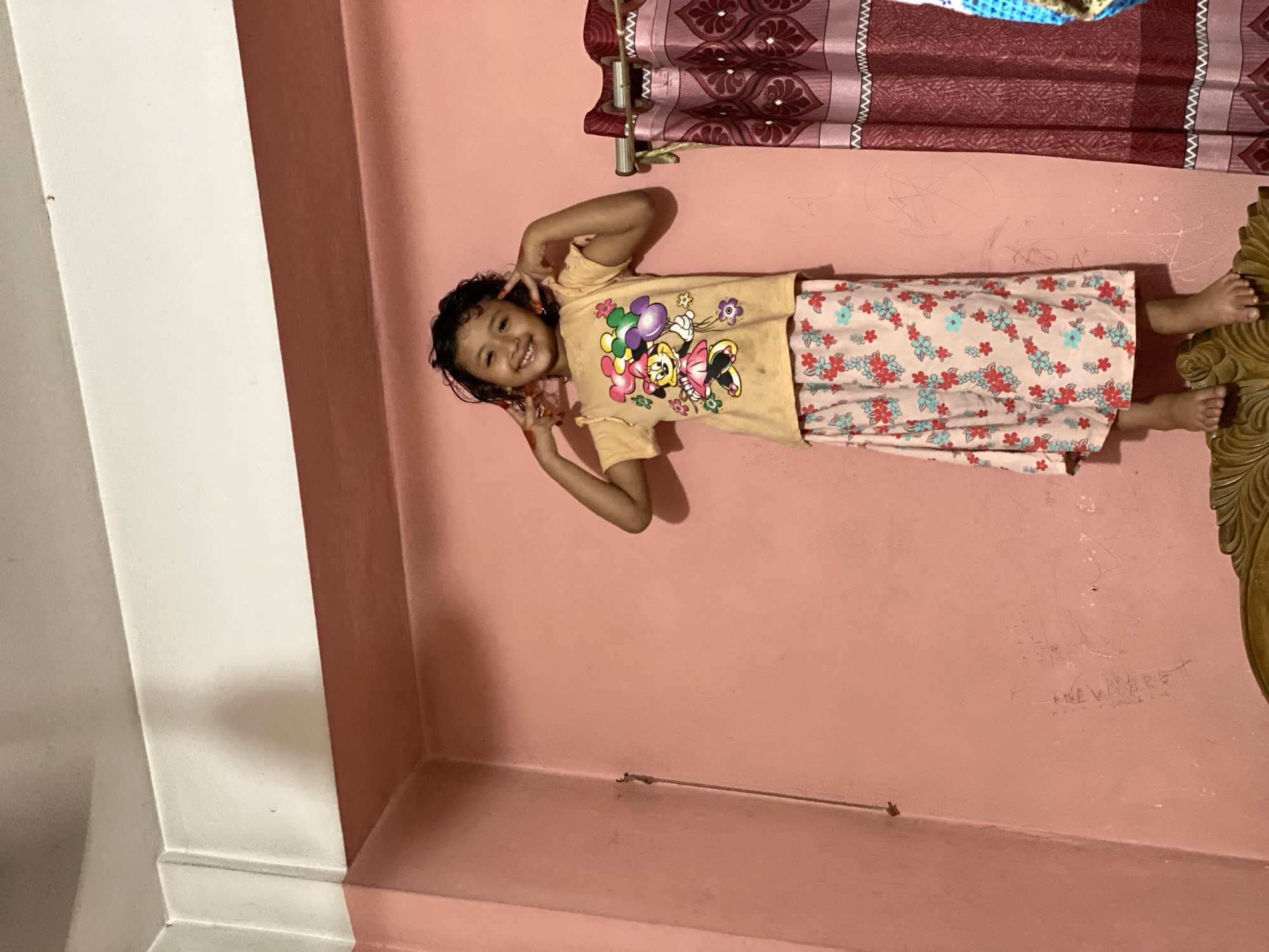 The image, originally rotated, depicts a young girl standing playfully indoors. She has dark, curly hair and a wide, joyful smile, making "peace" signs with both hands near her head. She is dressed in a light yellow t-shirt featuring colorful cartoon characters, including Minnie Mouse, and loose-fitting, floral-patterned pants or a skirt with a pink background adorned with red and light blue flowers.
The setting is a room, with a dominant light pink wall behind the girl and a white architectural column or wall section on the left. In the background, a patterned reddish-brown curtain hangs from a rod. The girl's bare feet are visible in the lower right, positioned near a piece of carved wooden furniture. A thin wire or cord runs horizontally along the lower portion of the pink wall. The wall shows some smudges and faint, scratched-on text, which appears to include partially legible letters that might spell "THE VALLERS." Traces of a reddish substance are visible on the girl's fingertips, possibly from play or an adornment. The scene conveys a cheerful and playful moment, likely captured in a domestic setting in Dhaka, Bangladesh. The lighting suggests an indoor environment.