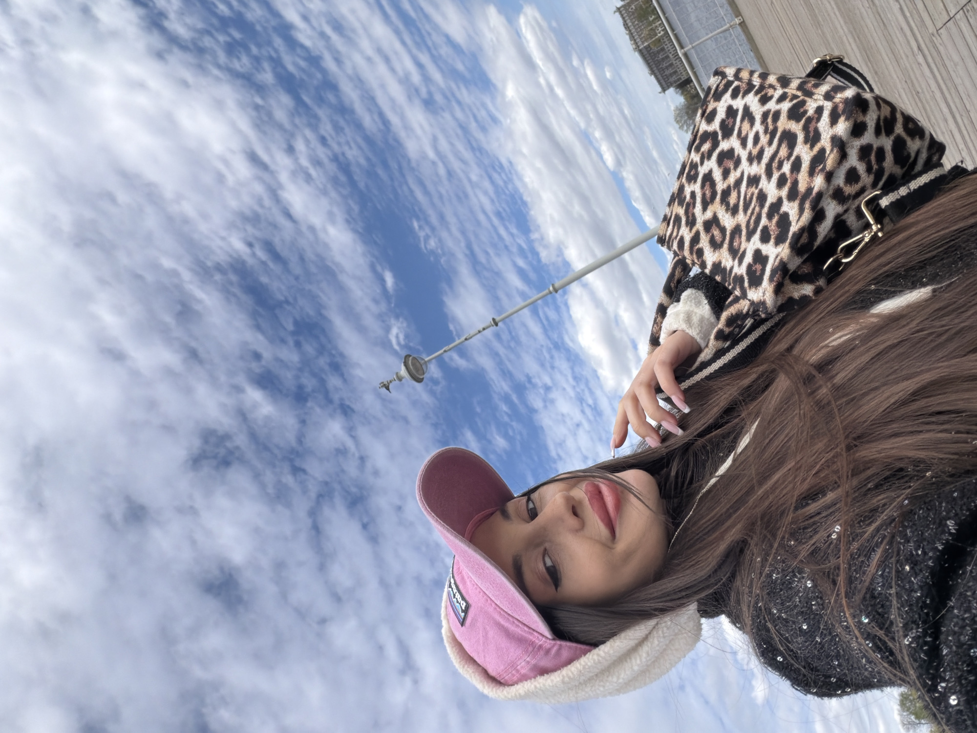 A woman wearing a pink cap and a leopard-print bag is posing for a photograph in Saint Petersburg, Russia. The background shows a cloudy sky and part of a building.