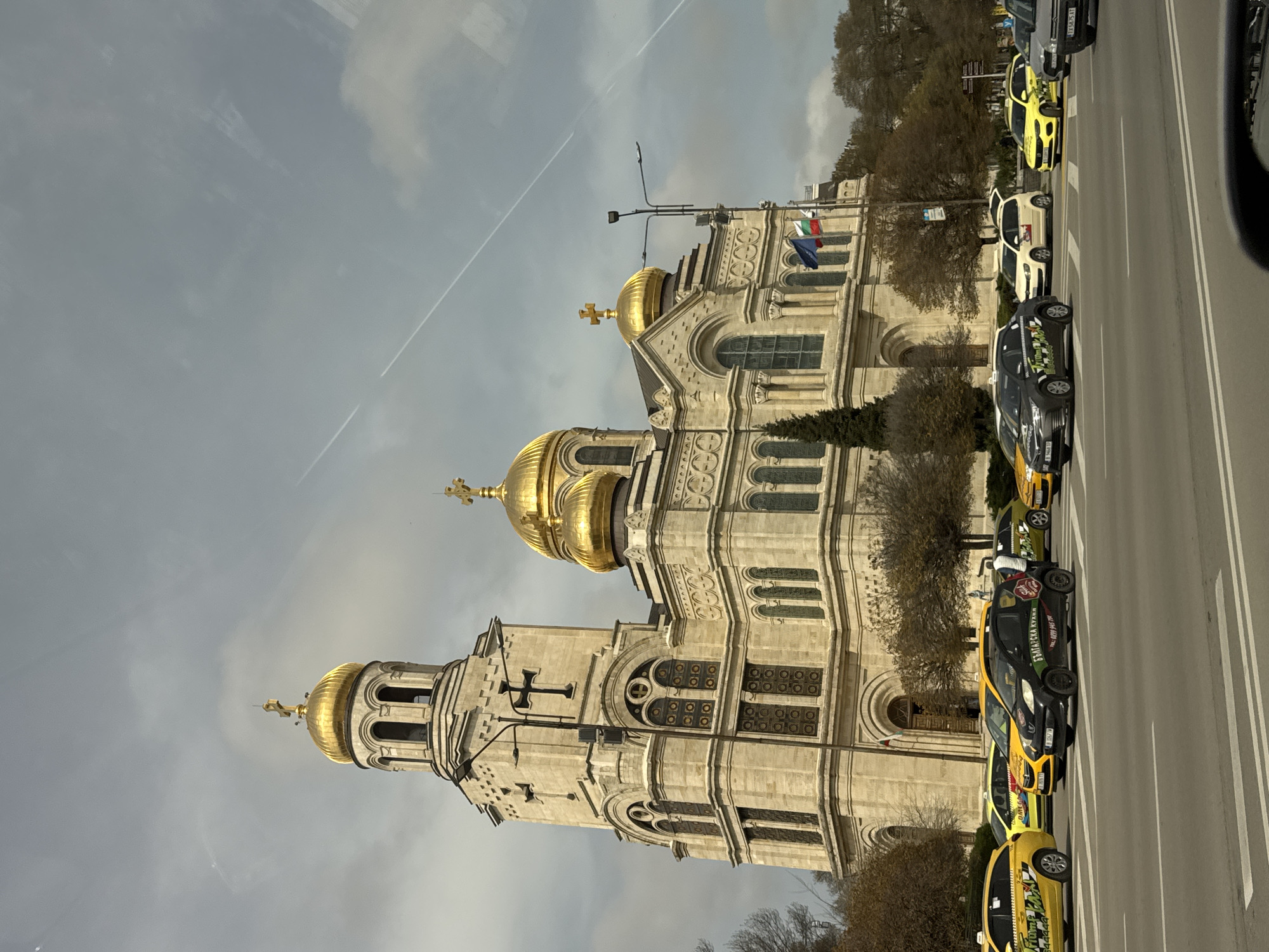 The image depicts the Dormition of the Mother of God Cathedral in Varna, Bulgaria, viewed from a street-level perspective. The architectural style of the cathedral is prominent, showcasing its golden domes and detailed facade. A row of taxis are parked along the side of the street next to the cathedral. The sky is overcast, with contrails visible. The Bulgarian flag is displayed near the top of the cathedral.