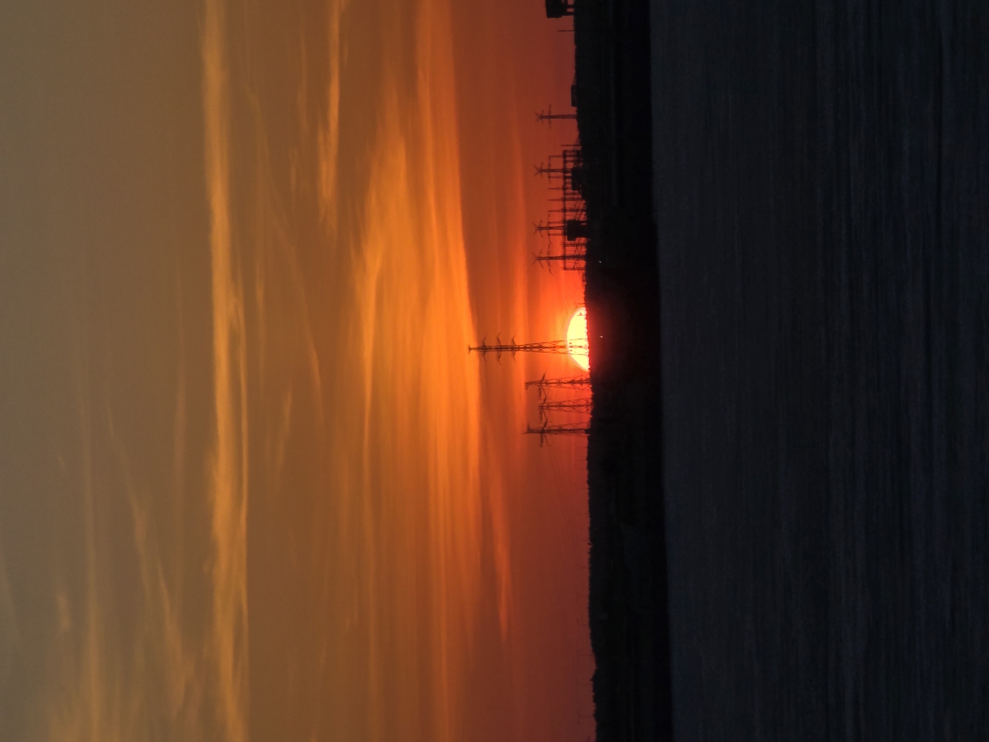 The image shows a sunset in Chaikovsky, Russia. The sun is setting behind power lines and some type of industrial plant, and the sky is filled with orange and yellow hues. A large body of water is visible to the right.