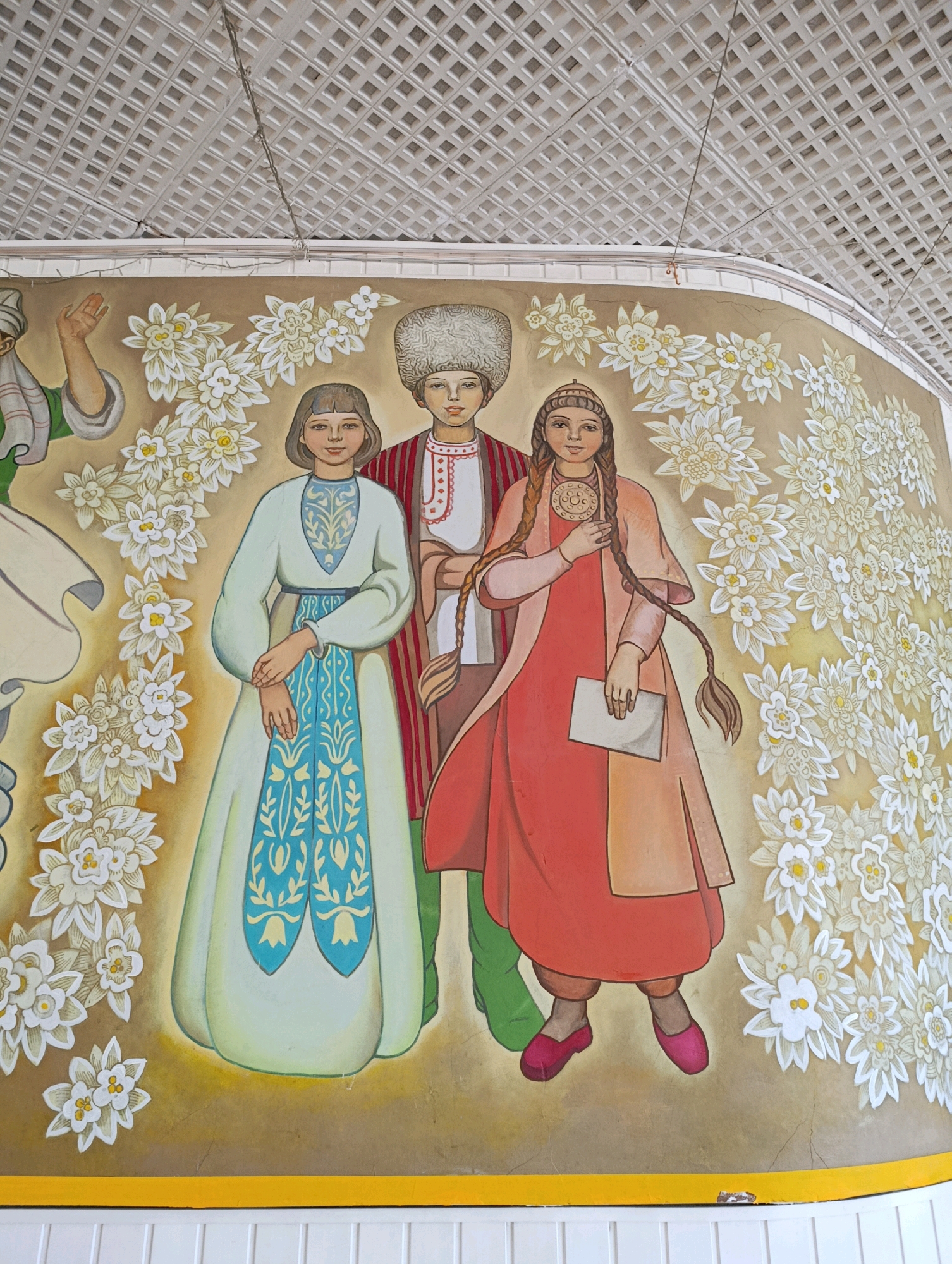 The image shows a mural on a curved wall. The mural depicts three people and floral motifs. The person on the left is wearing a headscarf and a green garment with a white and green striped sleeve. They are raising their right hand. To the right of this person is a girl wearing a light blue dress with intricate blue and yellow patterns on the bodice and skirt. She has short hair and her hands are clasped in front of her. In the center, a boy is wearing a large, furry hat, a white shirt with red embroidery, and a red and white striped robe. To the right of the boy is a girl with long braided hair. She is wearing a red dress and a light peach-colored overcoat. She holds a small piece of paper in her left hand and a round pendant hangs from her neck. All three figures are surrounded by stylized white and yellow flowers with green leaves. The background of the mural is a gradient of beige and yellow. The mural is located in Cherkasy, Ukraine, likely a public space given the presence of a suspended ceiling with a grid pattern above the mural, suggesting an indoor area such as a community center, a school, or a public building. A yellow border runs along the bottom of the mural, above white wooden paneling. There is a small area of damage on the yellow border near the bottom center of the image.