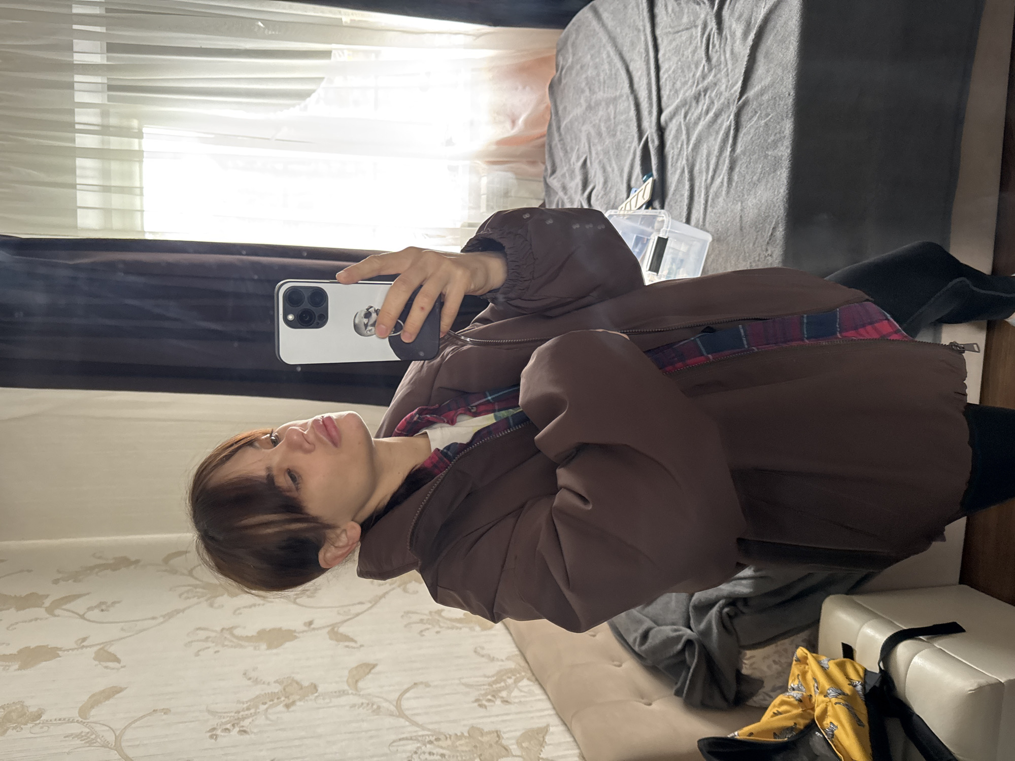 A person is holding a white smartphone with multiple rear cameras, possibly taking a selfie. The individual is wearing a brown jacket with a glimpse of a plaid shirt underneath. They appear to be in a room with floral wallpaper, a bed with gray and beige bedding, and a window covered by a sheer white curtain. There's additional clutter around the bed, including various items of clothing and bags. There is no definitive evidence to suggest this image was taken in Rostov-on-Don, Russia.