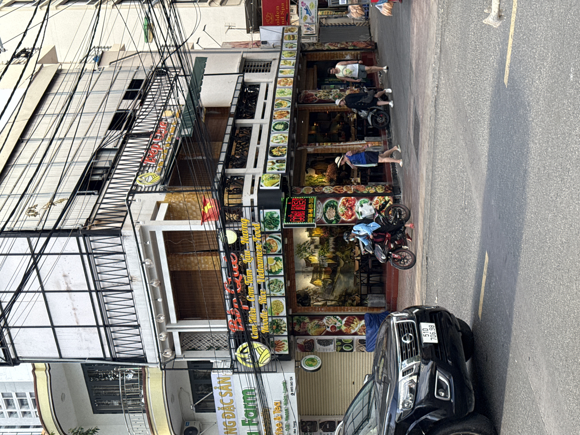 A street scene in Nha Trang, Vietnam, features a two-story building with a restaurant on the ground floor. The restaurant displays numerous photos of Vietnamese dishes along its facade, with the name "Búp Quế" visible on a sign. People are seen walking on the sidewalk in front of the restaurant, and a person on a motorcycle is stopped at the side of the road. A black pickup truck is parked on the left side of the image, with its license plate reading "51D 706.98". The building appears to be situated on a busy street, indicated by the presence of multiple vehicles and pedestrians. The architecture and signage suggest a vibrant urban environment typical of Vietnam.
