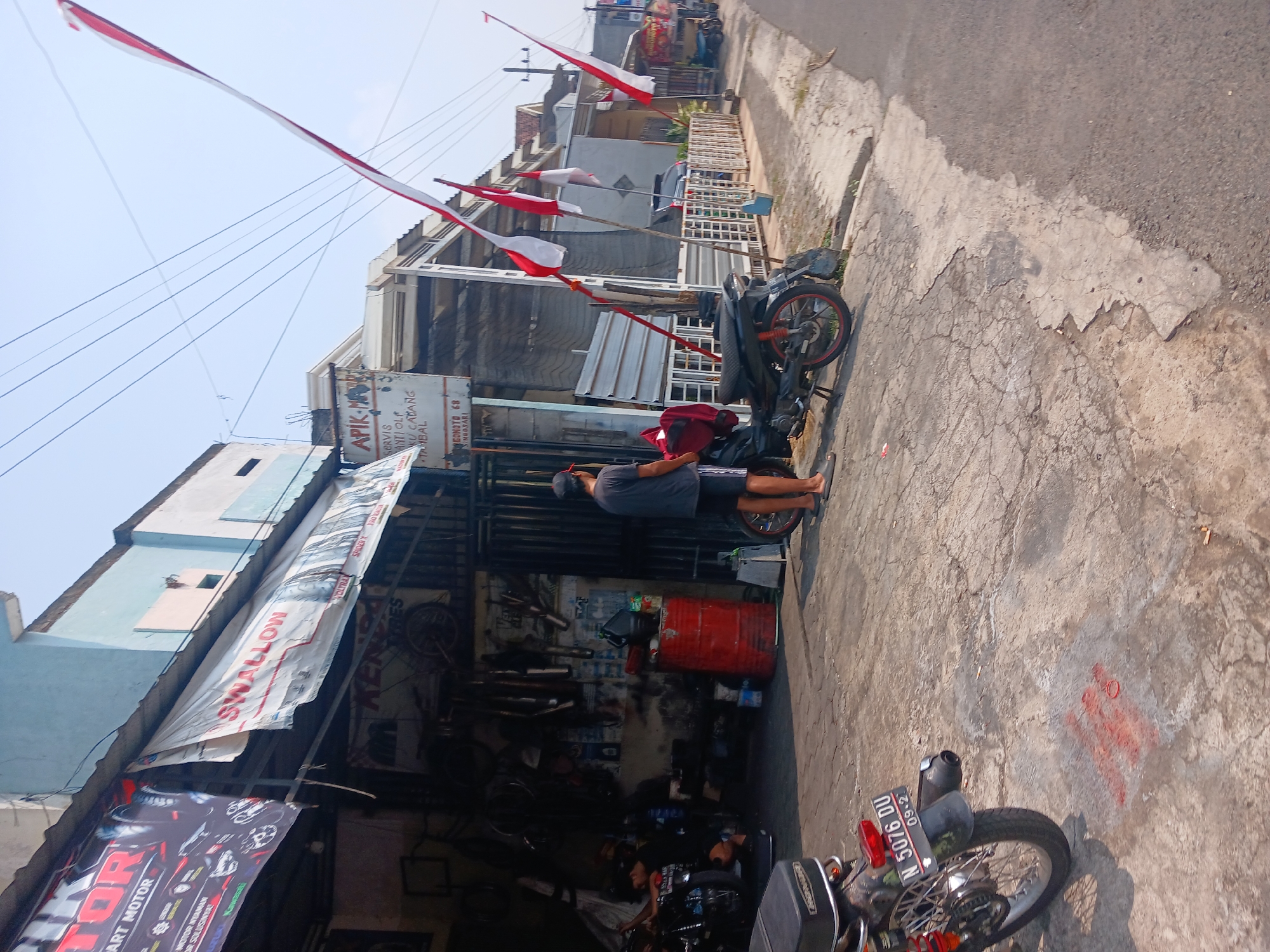 The image depicts a street scene in Singosari, Indonesia, likely an area with automotive repair shops. In the foreground, a dark-colored motorcycle with a license plate "N 5076 DU" is parked on a cracked concrete surface. To the left, another motorcycle is partially visible. In the background, a motorcycle repair shop is open, with tools and parts visible inside. A man in a dark t-shirt and shorts stands near the shop entrance. Red and white Indonesian flags are displayed prominently along the street. The buildings in the background appear to be part of the commercial establishment, with awnings and signs indicating services offered. The overall environment suggests a warm, sunny day.