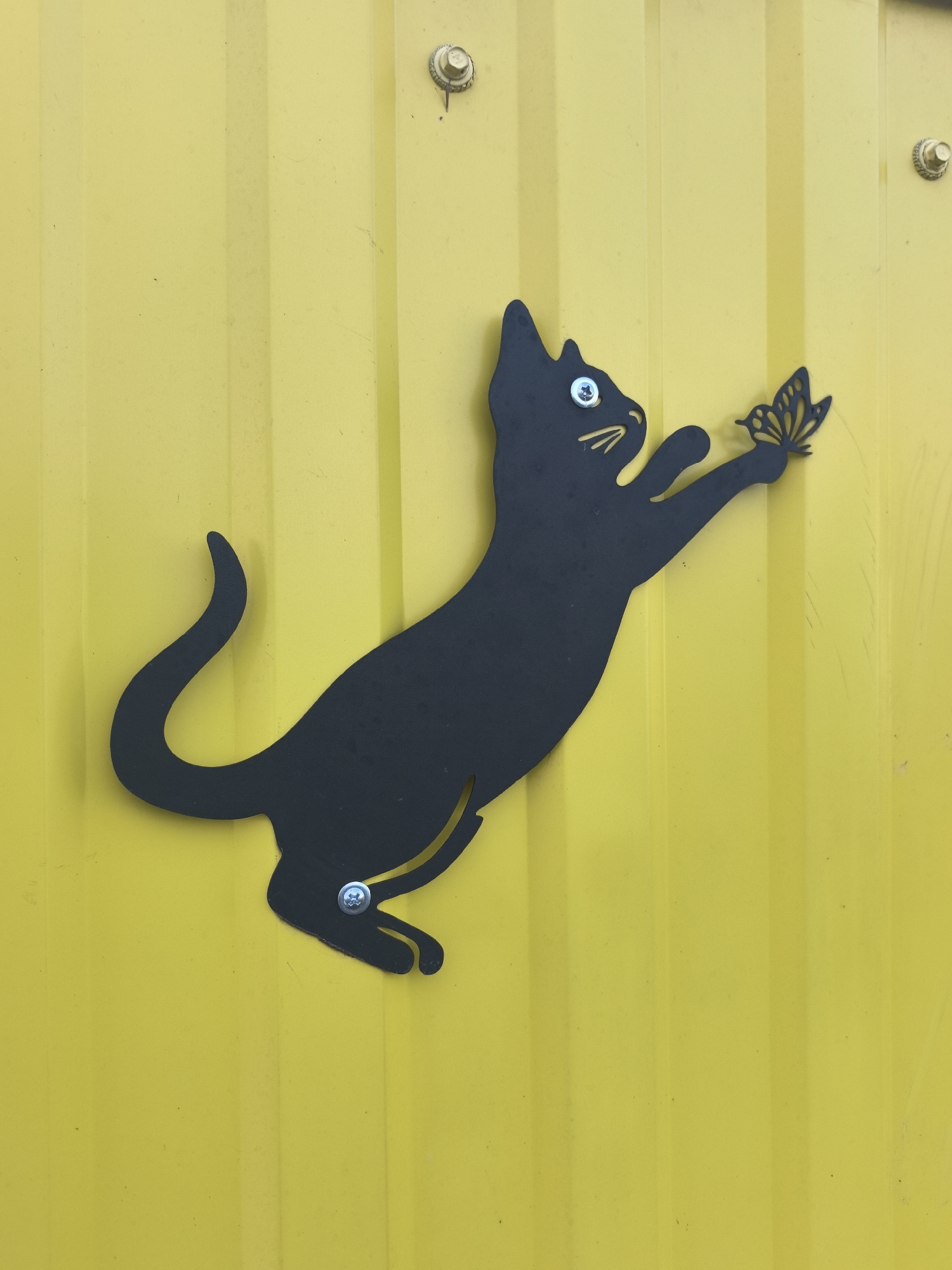 The image shows a black metal silhouette of a cat playfully reaching out to a butterfly, attached to a yellow metal wall with vertical ridges. The cat is mounted with screws in the eye and near the paws. There are other screws at the top of the wall. The location is not discernible from the image.