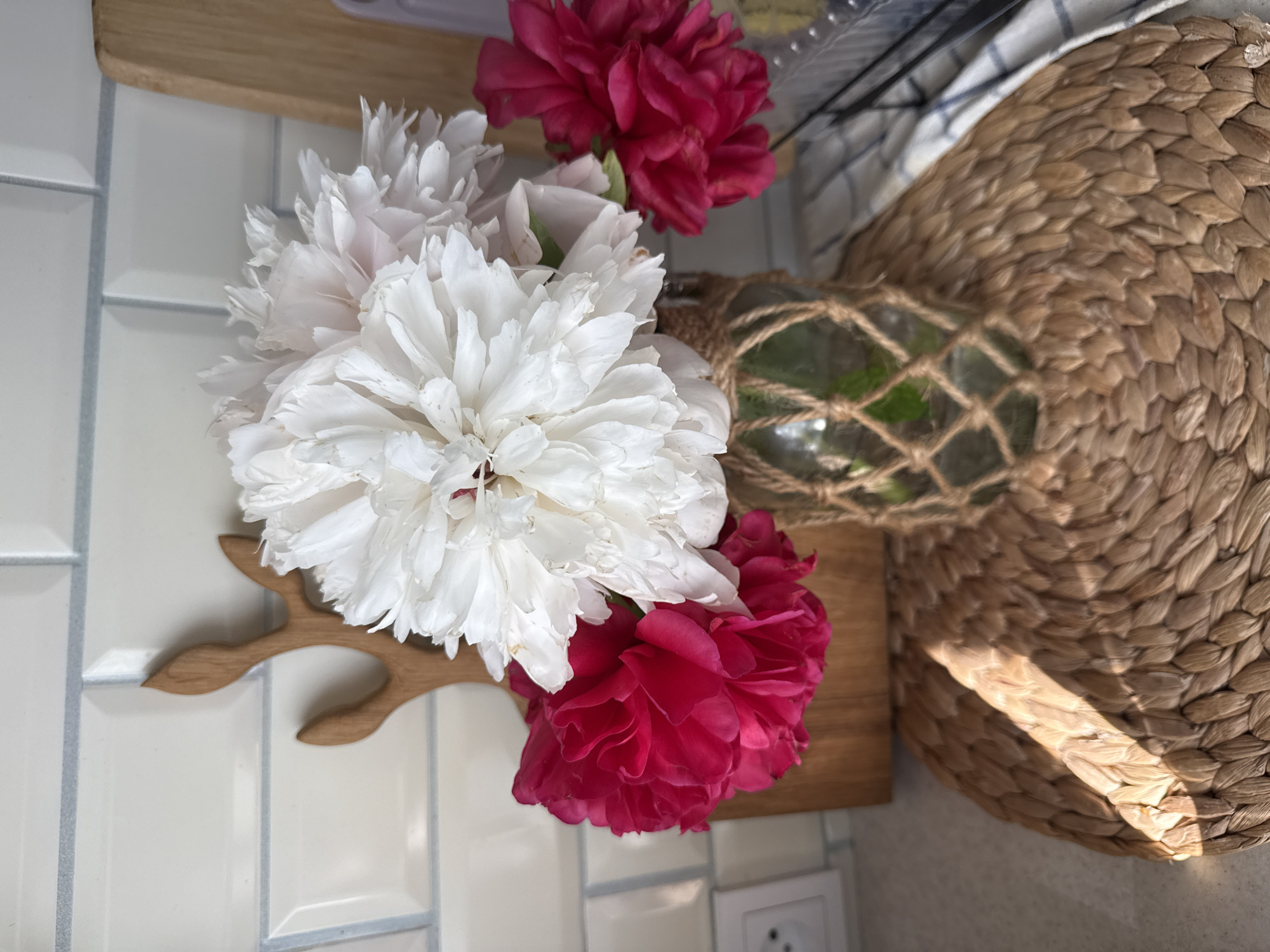 In a domestic setting, possibly a kitchen, a bouquet of peonies in a glass jar is displayed on a woven placemat. The jar is wrapped in twine, adding a rustic touch. Behind the flowers, a wooden cutting board with a deer antler design is propped against a tiled backsplash. The overall ambiance suggests a still life, capturing a moment of natural beauty within a home environment. There is no visible indication of the city of Stary Oskol, Russia.