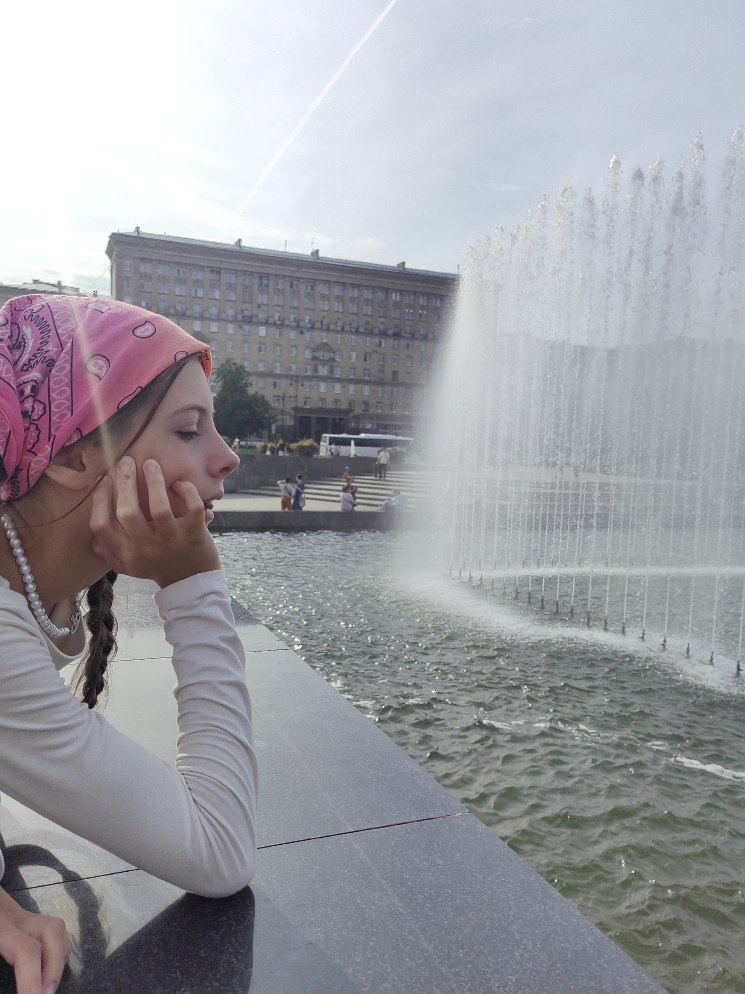 A young girl with a pink bandana and a pearl necklace looks out at a large fountain in Saint Petersburg, Russia. She is wearing a long-sleeved white shirt and has her hand on her chin. Her hair is in two braids. The fountain is large and impressive, with water spraying upwards in a curtain-like fashion. In the background, a large building with many windows can be seen, as well as several people walking around. The sky is bright and clear, with a contrail visible. The water in the fountain is rippling and reflects the sunlight. The girl is seated on a dark, polished surface, likely a bench or ledge, with the fountain's water in front of her.