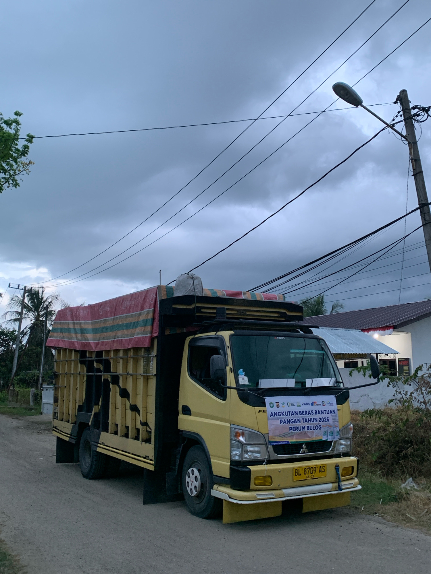 A yellow truck with a tarp-covered cargo area is parked on a dirt road in what appears to be Pantegurah, Indonesia. The truck has a banner on its front that reads "ANGKUTAN BERAS BANTUAN PANGAN TAHUN 2026 PERUM BULOG." This translates to "Rice Transportation Aid Food Year 2026 Perum Bulog," suggesting it's involved in food aid distribution. The sky is overcast, with visible power lines and a street lamp overhead. In the background, there are houses and trees, with palm trees also visible. The overall scene depicts a rural or semi-rural setting.