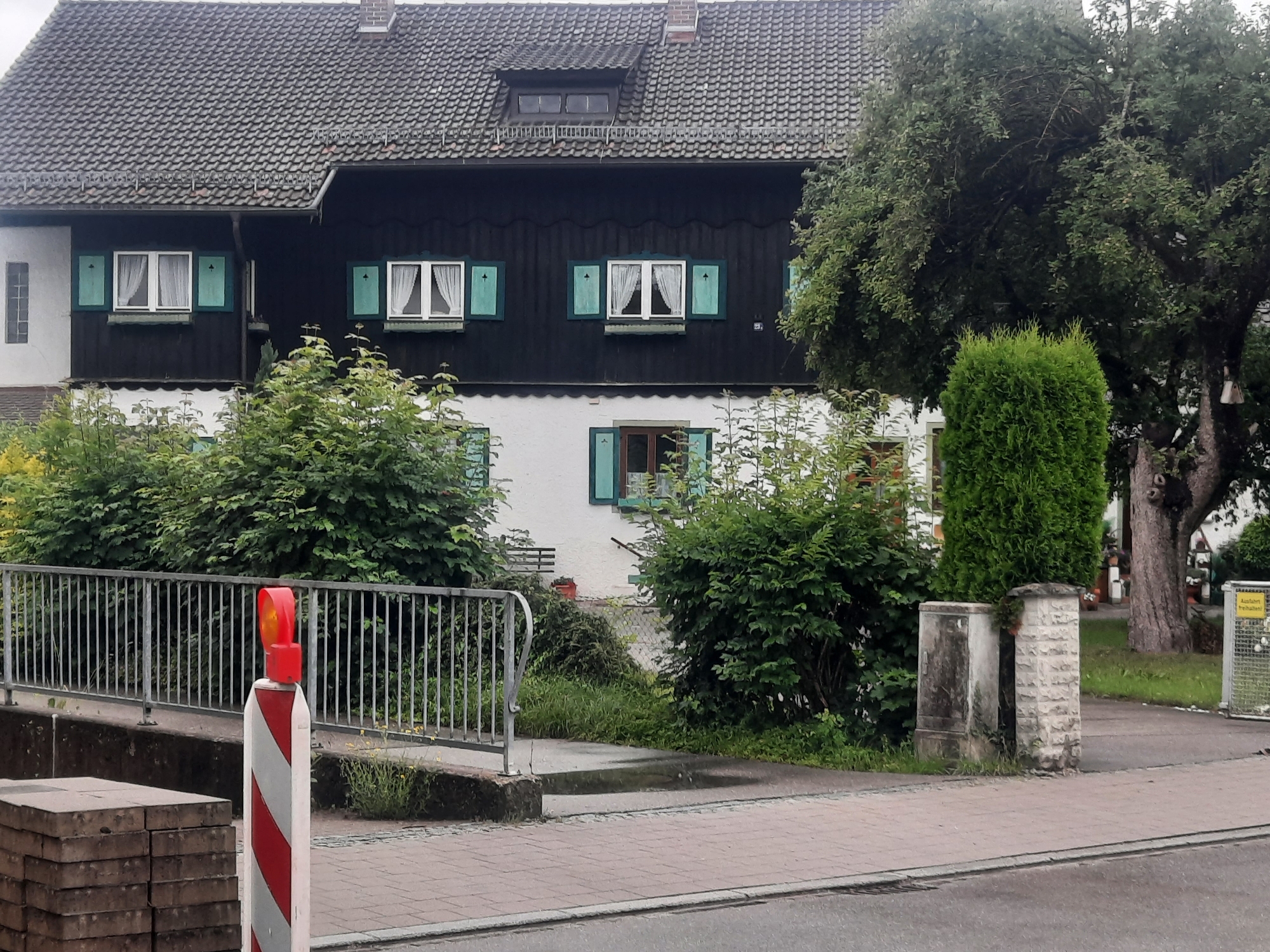 The image shows a street-level view of a two-story house with a dark, wood-paneled upper facade and a white-painted lower facade. The roof is covered in dark, grey tiles and features a dormer window. The house has several windows with light-colored curtains and green shutters. To the right of the house, a large, leafy tree dominates the scene. In the foreground, a metal railing lines a walkway, and a red and white striped traffic post with a flashing orange light is visible. Paving stones and a section of asphalt road are also present. Green bushes and some overgrown grass are scattered throughout the scene.