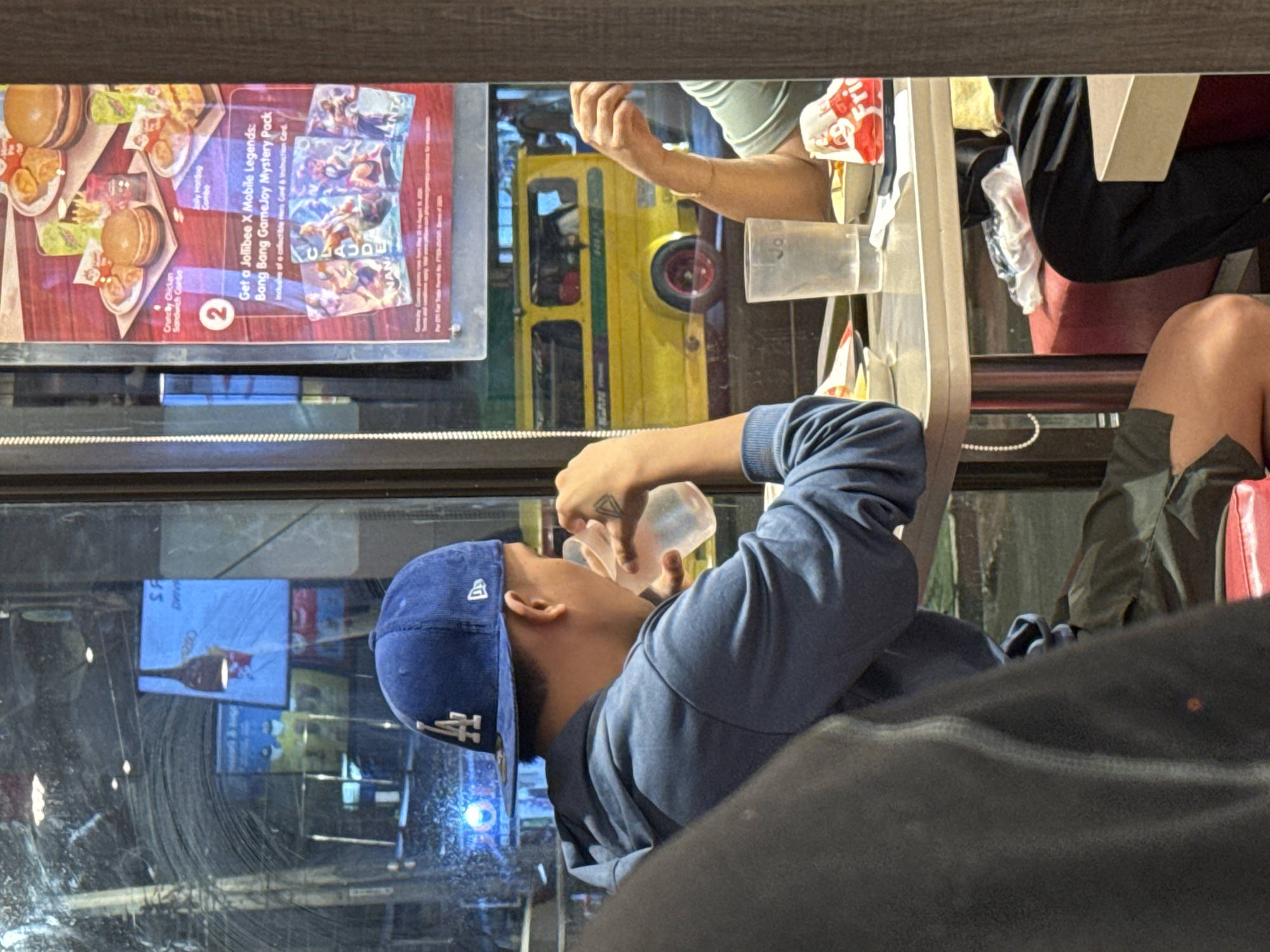 The image presents an indoor scene, likely within a Jollibee restaurant in Davao City, Philippines, viewed through a large window. The central subject is a person, seen in profile, wearing a blue long-sleeved top and a blue baseball cap with a white "LA" logo. They are in the act of drinking from a clear plastic bottle, holding it to their mouth. A small, dark triangular tattoo is visible on the back of their right hand.
The window serves as a reflective surface, showing elements from both inside and outside. To the left, a Jollibee advertisement is prominently displayed, showcasing food items like burgers and hotdogs. The ad promotes a "Jollibee X Mobile Legends: Bang Bang GameJoy Mystery Pack," which includes a "collectible Hero Card & Instruction Card," with hero names like "CLAUDE" and "NAN" visible on the game cards. The ad also mentions "Crunchy Chicken Sandwich Combo" and "Jolly Hotdog Combo," along with partial text like "SA," "DHIVA," "2," "Jo," and "Fri."
Reflected outside the window is a distinct yellow jeepney with red wheel hubs, a characteristic form of transportation in the Philippines. Other reflections include what appears to be a blue advertising screen or sign. Inside the restaurant, parts of other individuals are visible, including an arm in a light green sleeve, and legs, one clad in dark fabric, resting on a red seat. General restaurant elements like red seating and plastic containers are also present. The lighting indicates daytime, with bright outdoor light contributing to the window's reflections.