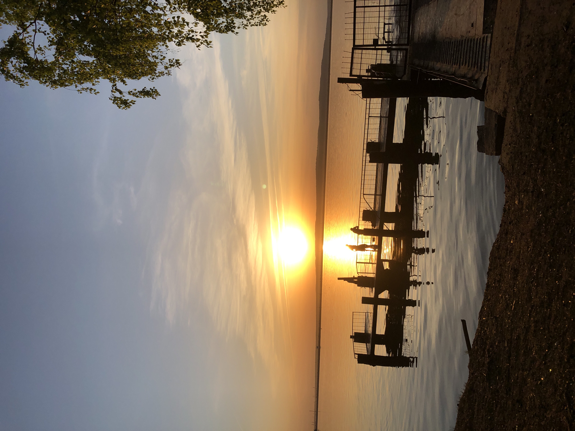 The image shows a sunset scene, likely taken in Chaikovsky, Russia, on the Kama River. The sun is setting over a body of water, casting a strong golden glow. In the middle ground, there is a silhouette of a metal structure, possibly a pier or dock. The silhouette suggests the presence of people on the structure. To the left of the image, branches of a tree are visible. The foreground shows the edge of the shore, possibly gravel or soil. The overall environment is serene and focused on the sunset.
