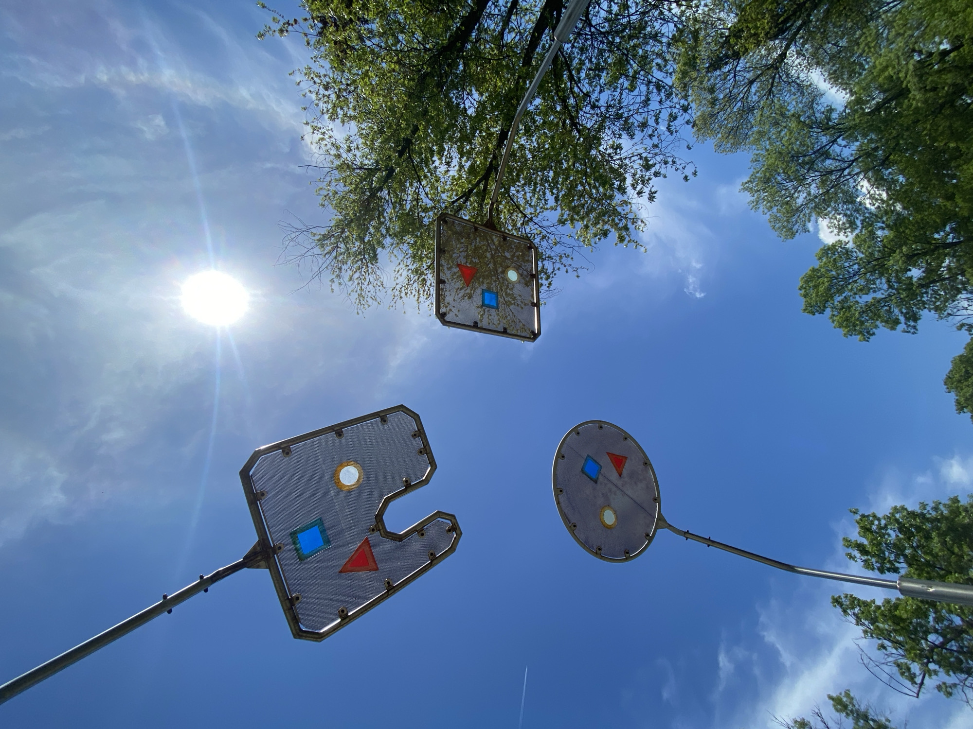 Looking up at the blue sky with the sun shining brightly, there are three metal structures on poles, seemingly part of an outdoor art installation. The structures have geometric shapes on them: squares, circles, and triangles, some filled with blue or red. These metal structures are partially framed by green tree branches. The photo was likely taken at a children's playground in the city of Sofia, Bulgaria.