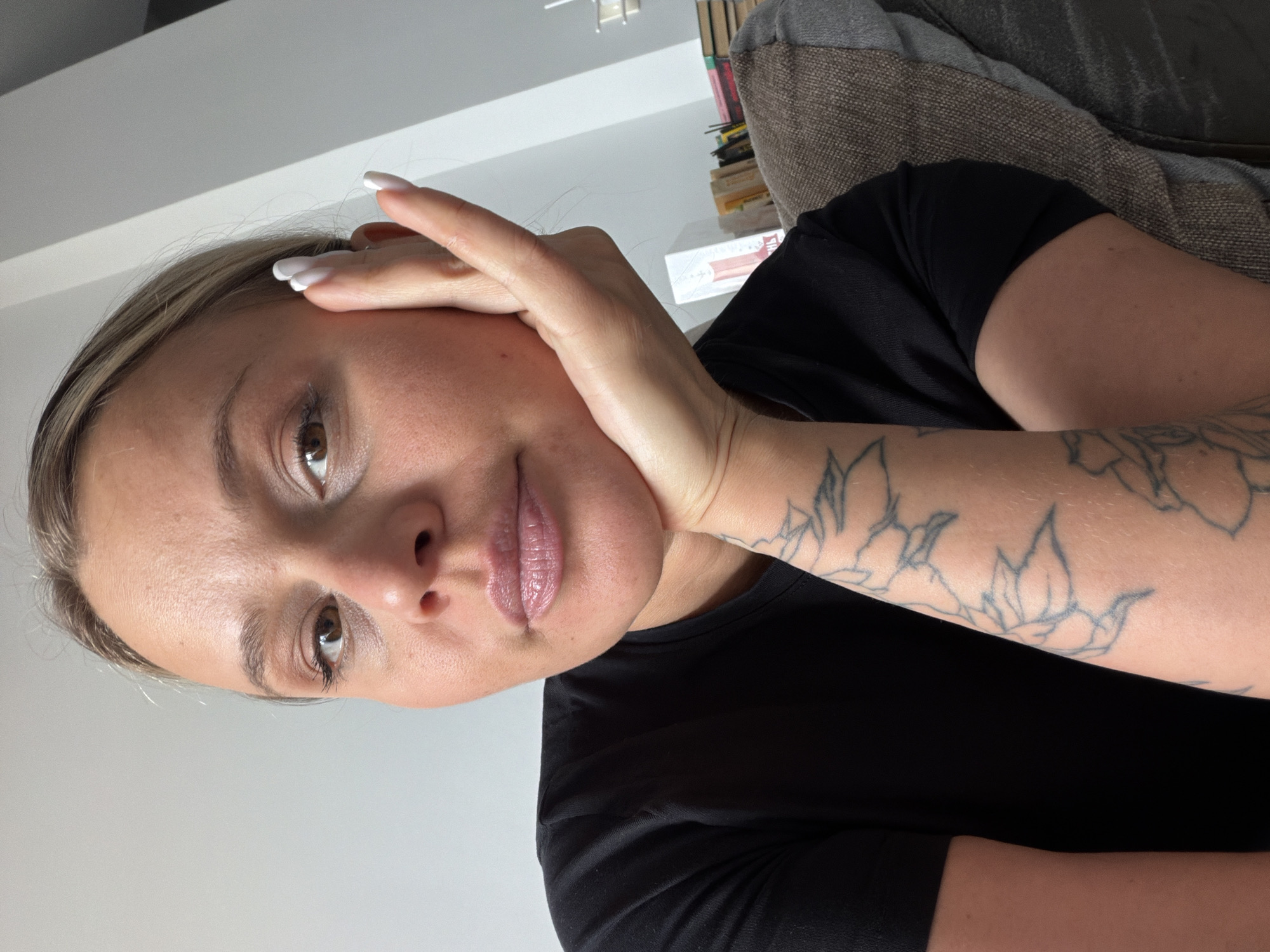 A woman with light brown eyes and fair skin is looking at the camera with a slight smile. She is wearing a black shirt. Her left hand is supporting her face. She has a tattoo of flowers on her left forearm. The background includes a light gray wall, a stack of books, and a gray couch. The location is not evident in the image.