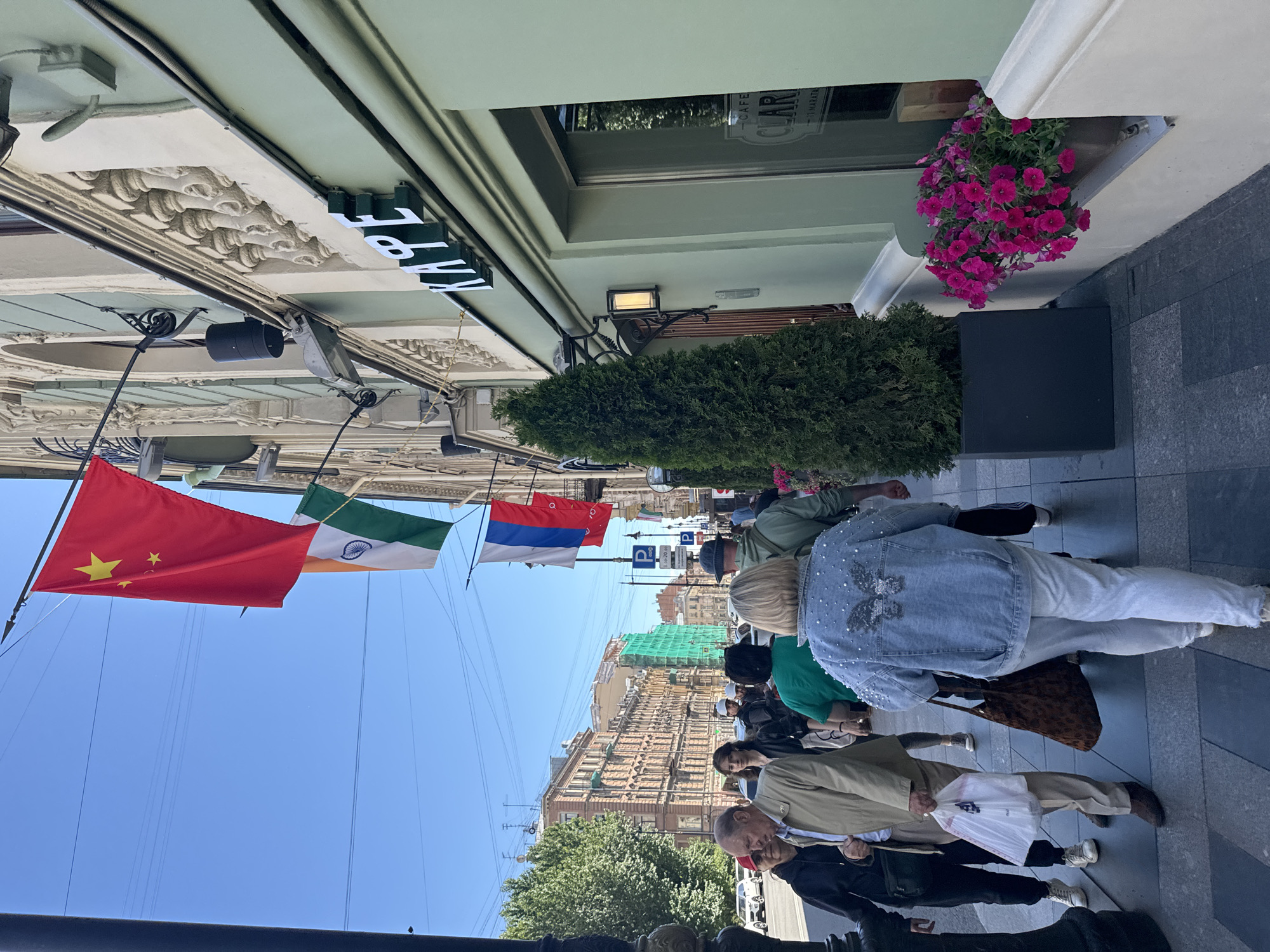 The image depicts a sunny day on a city street in Saint Petersburg, Russia, likely a pedestrian area. In the foreground, several people are walking on a sidewalk. A woman in a denim jacket adorned with pearls and a butterfly motif is prominent, walking away from the camera, followed by other pedestrians. To the left, an older man and a woman are walking towards the camera. The building on the right has a light green facade and features a sign with the word "КАФЕ" (KAFE, meaning cafe) in white letters. The building's architecture includes ornate decorative elements along the upper facade. Flags are displayed above the street, including the flag of China, the flag of India, and the flag of Russia. There are also trees in planters and what appear to be parked cars in the background, along with the facade of a multi-story building. The overall scene suggests a typical urban streetscape with commercial establishments and pedestrian activity.