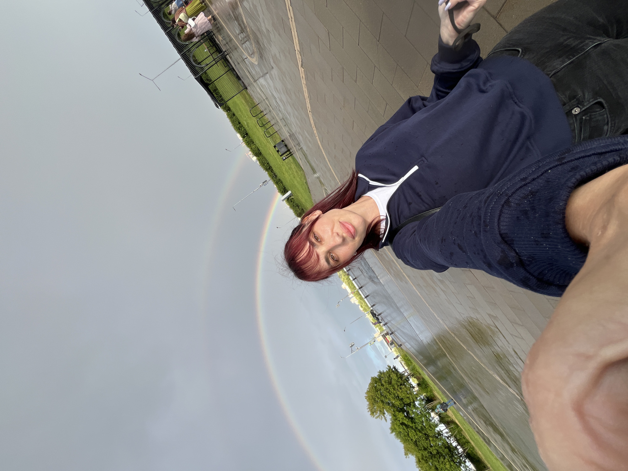 This is a selfie taken outdoors, with the camera tilted. The primary subject is a young woman with reddish-brown hair, wearing a navy blue hooded sweatshirt over a white t-shirt and dark jeans. She is looking at the camera with a slight smile. In the background, a double rainbow is visible in a cloudy, gray sky. The ground appears wet, suggesting recent rain. A paved walkway runs diagonally across the frame, with a grassy area to the left. There are some trees and distant structures visible on the right side of the image. The scene appears to be a public outdoor space, possibly a park or promenade, in Nizhny Novgorod, Russia. The overall mood is casual and pleasant, despite the rainy weather that likely caused the rainbow. No visible text is present in the image.