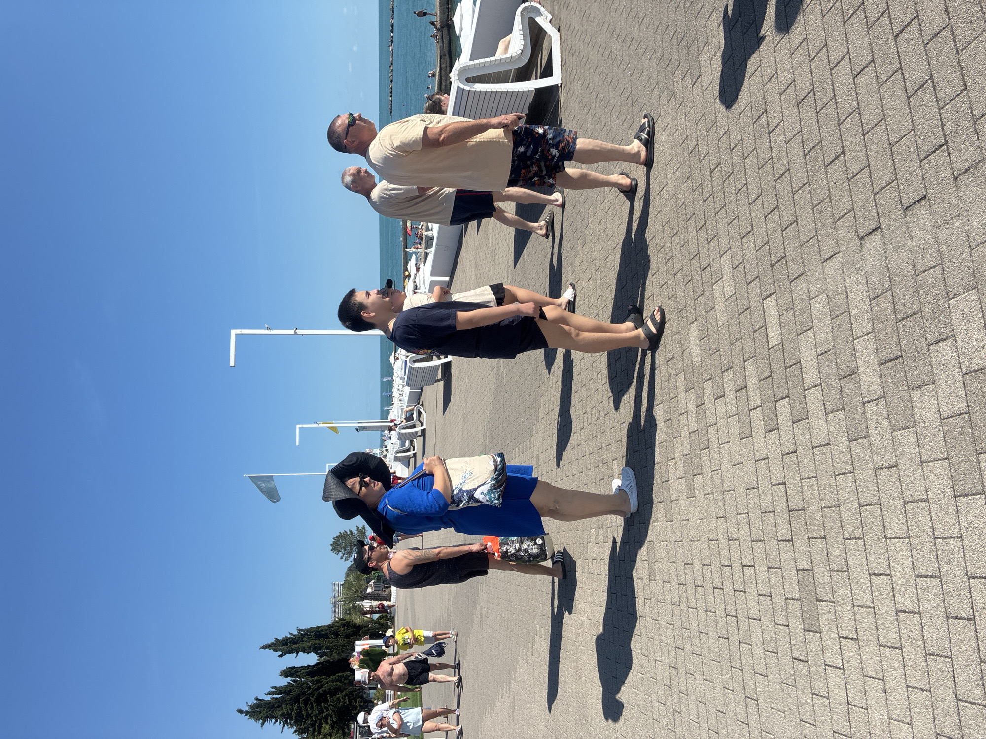The image depicts a sunny day on a paved promenade adjacent to the sea. In the foreground, a group of people are walking and standing. A woman in a blue dress and a large black hat is prominent, carrying a tote bag with a Japanese wave print. To her left, a man in a black tank top and shorts holds a patterned bag. Further down the promenade, several men in casual summer attire are visible, some walking and others standing. Beyond the promenade, white benches and umbrellas line the area near the sea, and in the distance, people are visible by the water. The sea itself is calm and blue, with a rocky breakwater in the middle distance. The sky is clear and bright blue. The architectural style of the buildings and the overall atmosphere suggest a resort location. Based on the context of Sochi, Russia, this scene likely represents a typical day on one of its many seaside promenades.