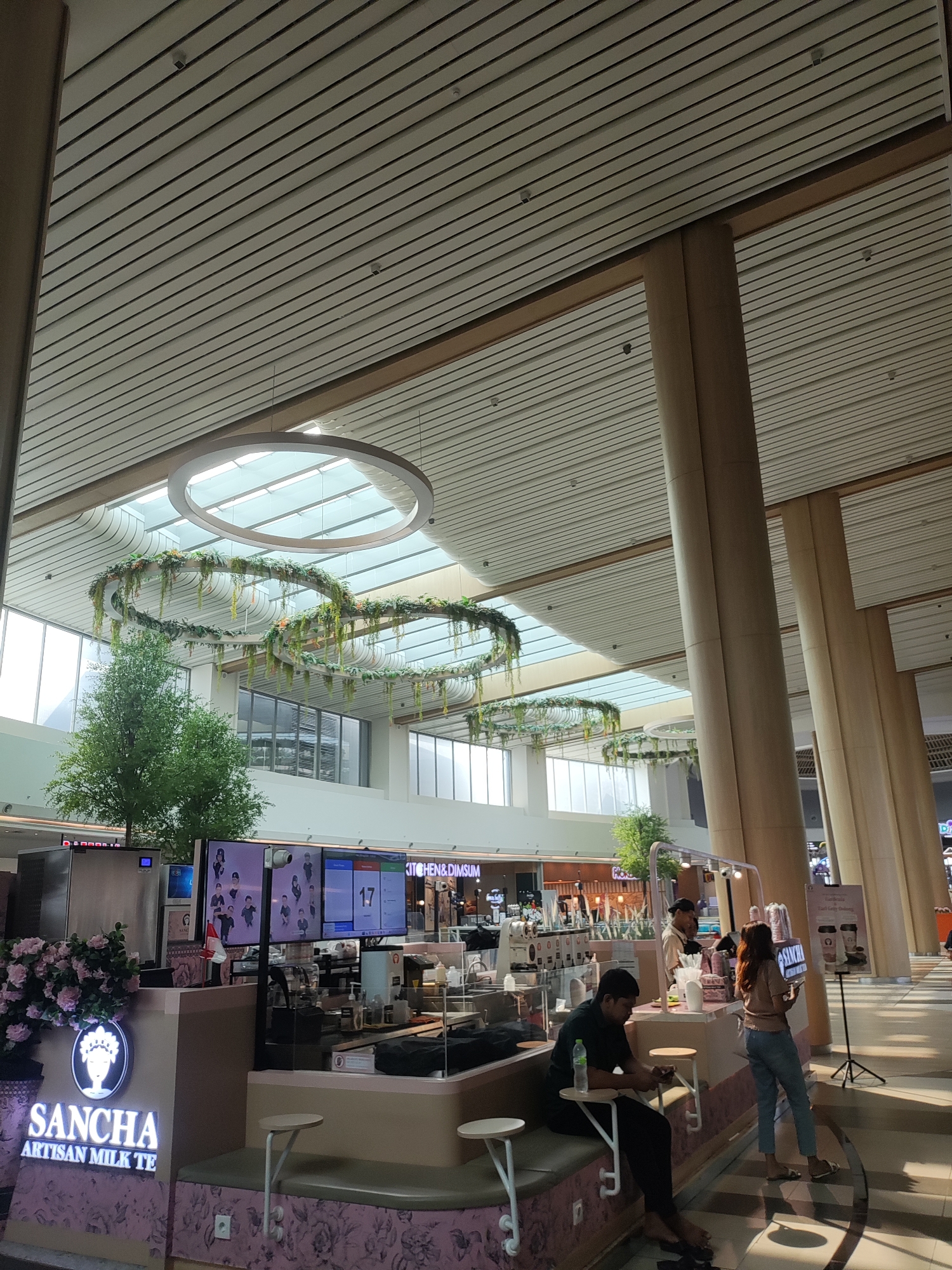 The image depicts a contemporary indoor space, possibly a shopping mall or airport terminal, with a high, slatted ceiling and large pillars. Decorative circular structures adorned with greenery hang suspended from the ceiling. In the foreground, a "SANCHA Artisan Milk Tea" stall is visible, featuring a pink and white counter with a seating area. Several individuals are present: one person is sitting at the counter looking at their phone, another person is standing and appears to be a customer or staff member at the stall, and two individuals are visible behind the counter, one of whom is a staff member. A digital display board behind the counter shows what appears to be a queue number and images of people. The overall atmosphere suggests a commercial or public waiting area. The location is identified as Lambangjaya, Indonesia, but this information is not visually verifiable from the image itself.
