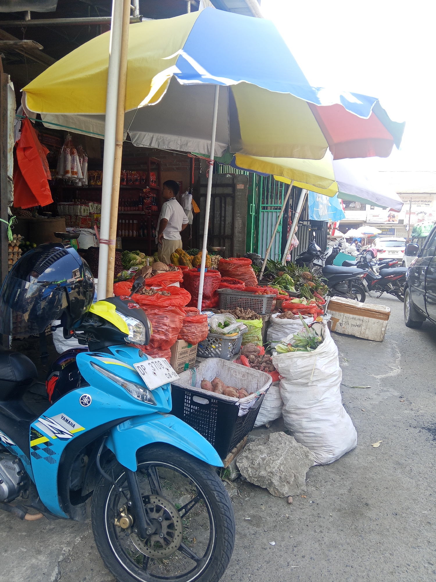 The image depicts an outdoor market scene in Makale, Indonesia, on a bright day. In the foreground, a blue Yamaha motorcycle is parked, equipped with a black helmet on its handlebars and a yellow accent on its front. The motorcycle has a license plate that reads "DP 3708 JB 08-25".
Behind the motorcycle, a market stall is set up under large, colorful umbrellas (yellow, blue, and red). The stall displays a variety of goods, predominantly fresh produce. Numerous red plastic bags filled with items, likely chili peppers or similar vegetables, are visible. There are also sacks and baskets containing what appear to be root vegetables and greens, including what looks like pineapples. A young boy in a white shirt stands near the stall, observing the surroundings.
The market is situated on a paved street, with other motorcycles and a dark-colored vehicle parked in the background, suggesting an active commercial area. The overall environment is that of a bustling local market, characteristic of a town in Indonesia like Makale.